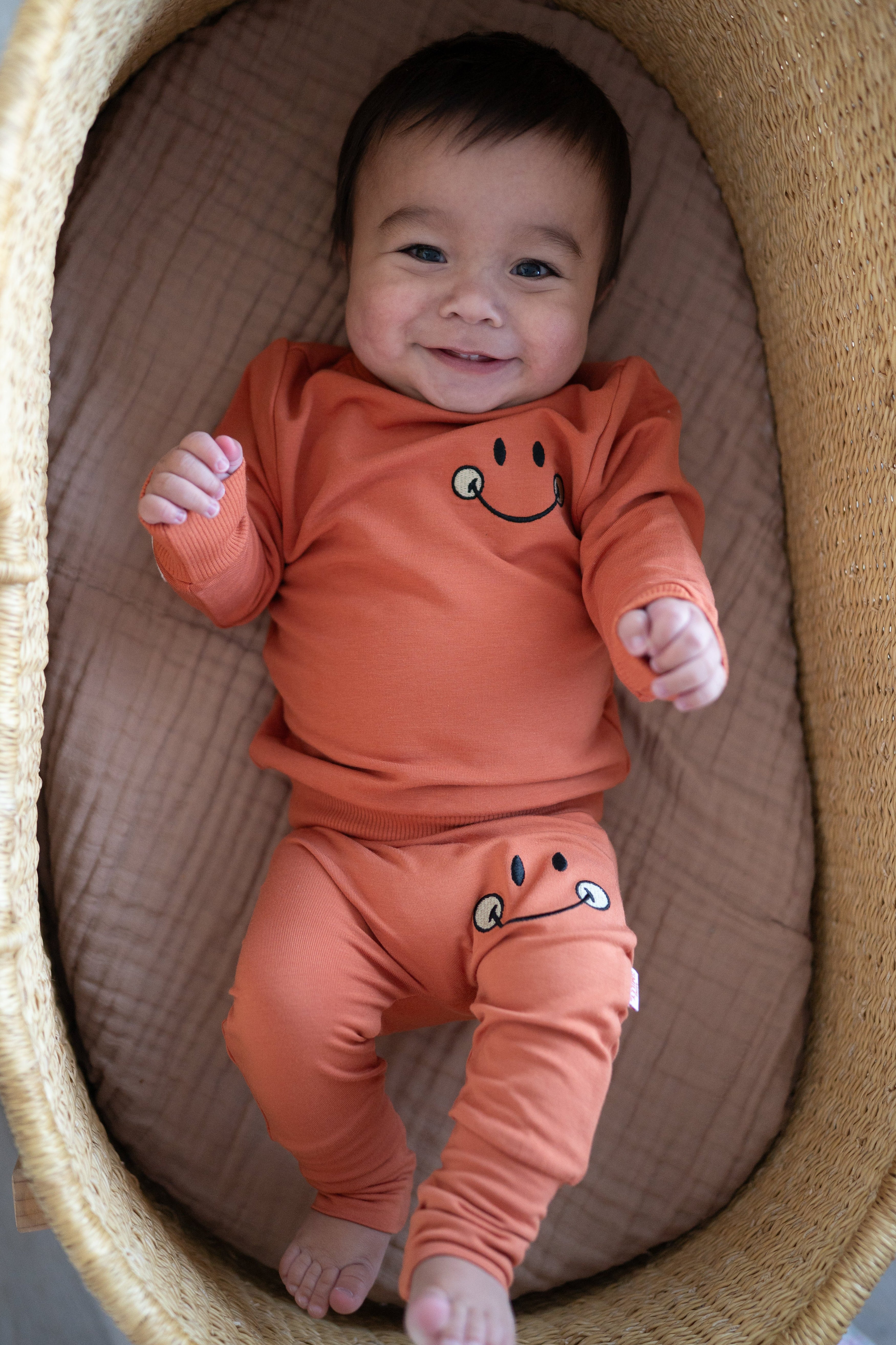 A baby wearing a rust-colored crewneck shirt and pants with a smiley face design.