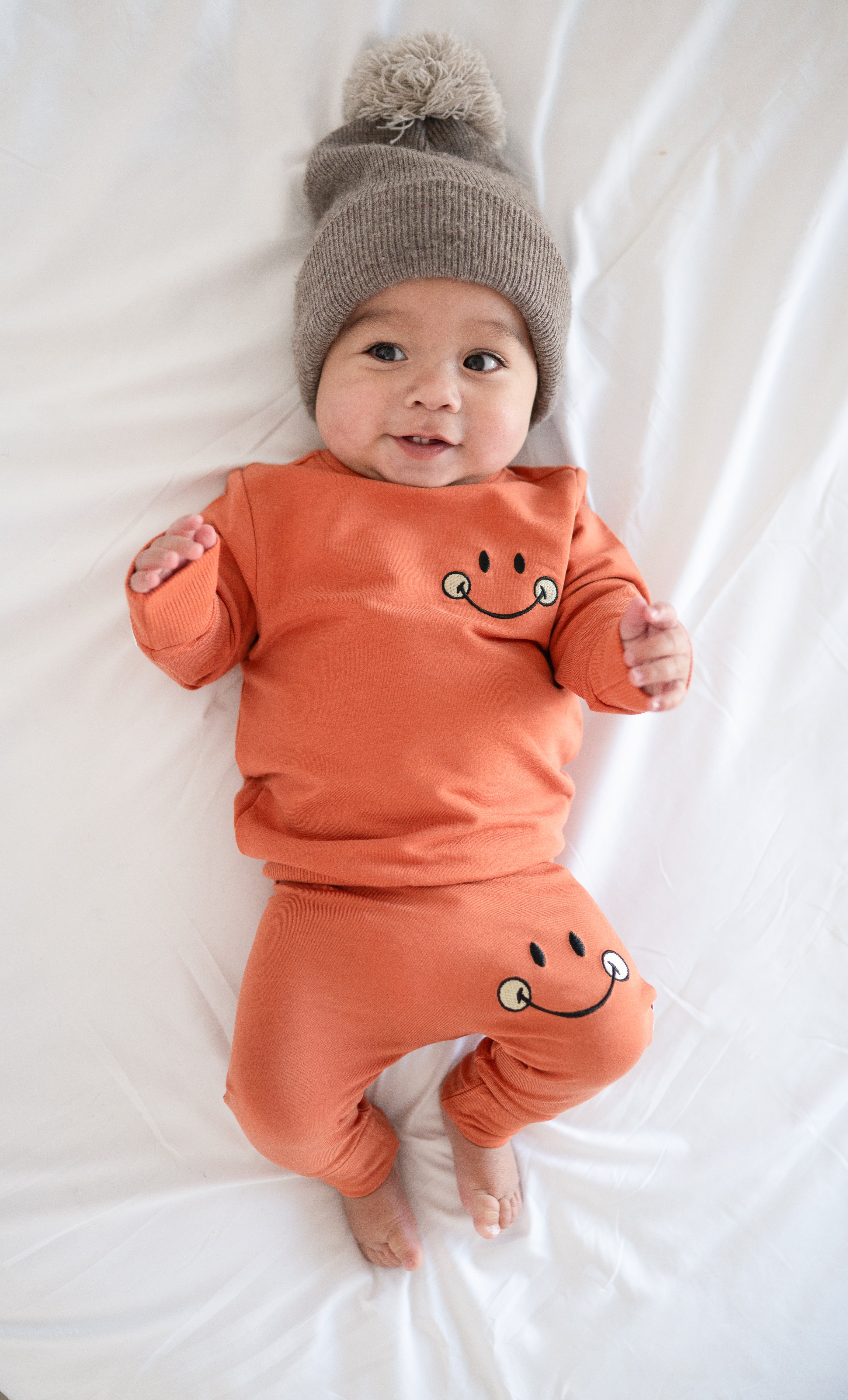 Baby in orange pants and sweatshirt with smiley faces, wearing a gray beanie.