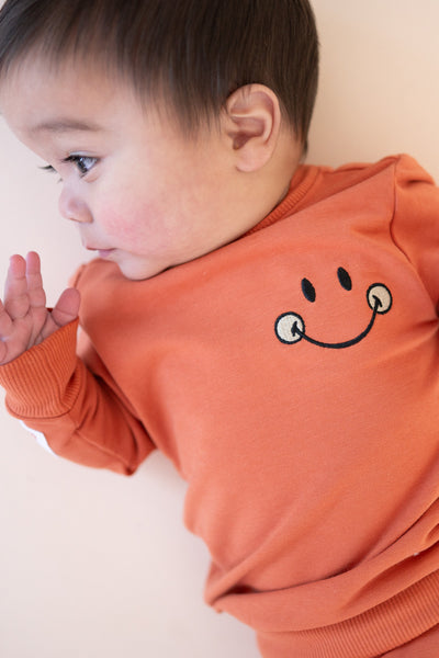 A baby wearing an orange crewneck sweatshirt with an embroidered smiley face.