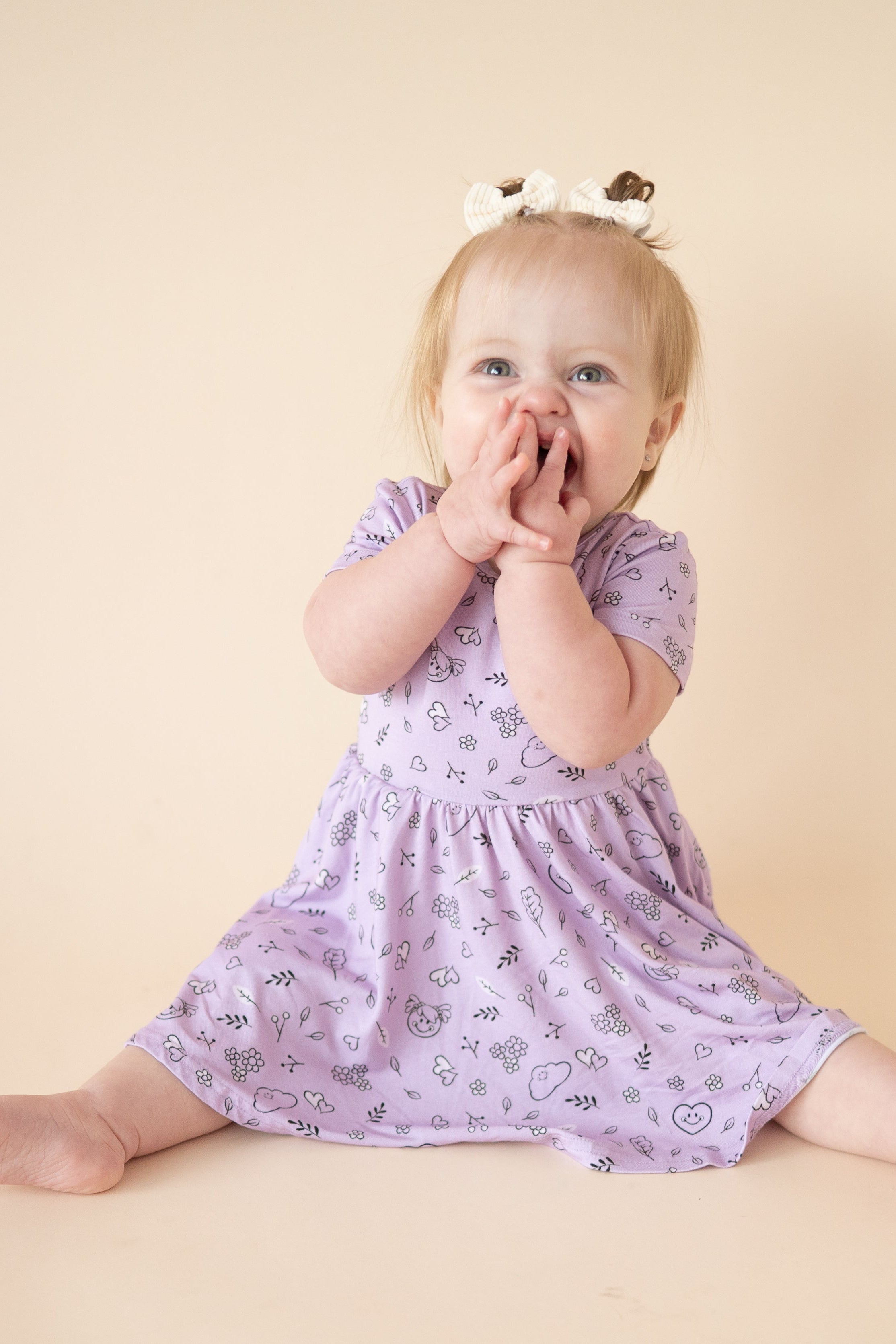 A baby girl in a lilac dress with a doodle print sits with her hands over her mouth.