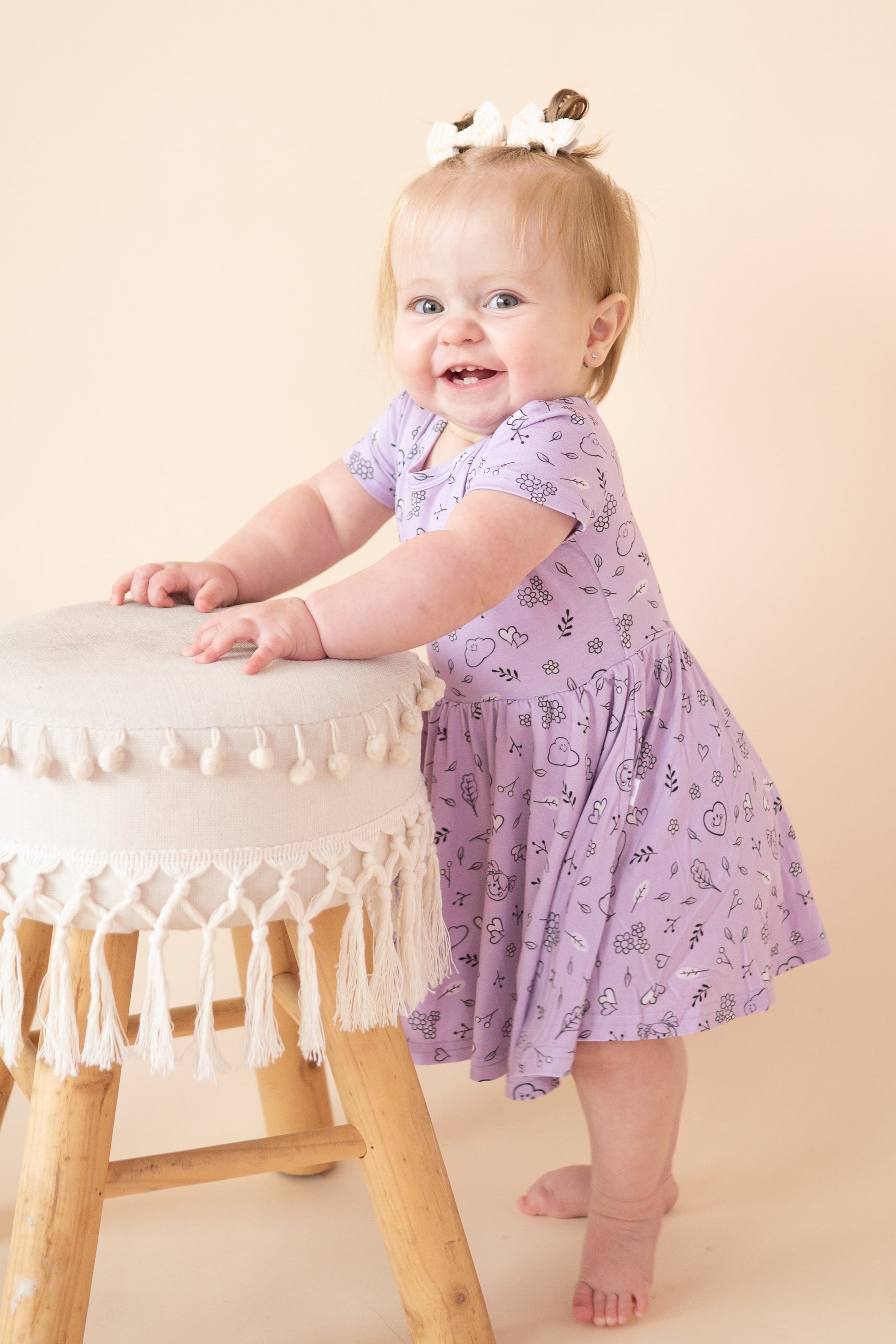 Baby in a lilac dress with a doodle print, standing by a stool.