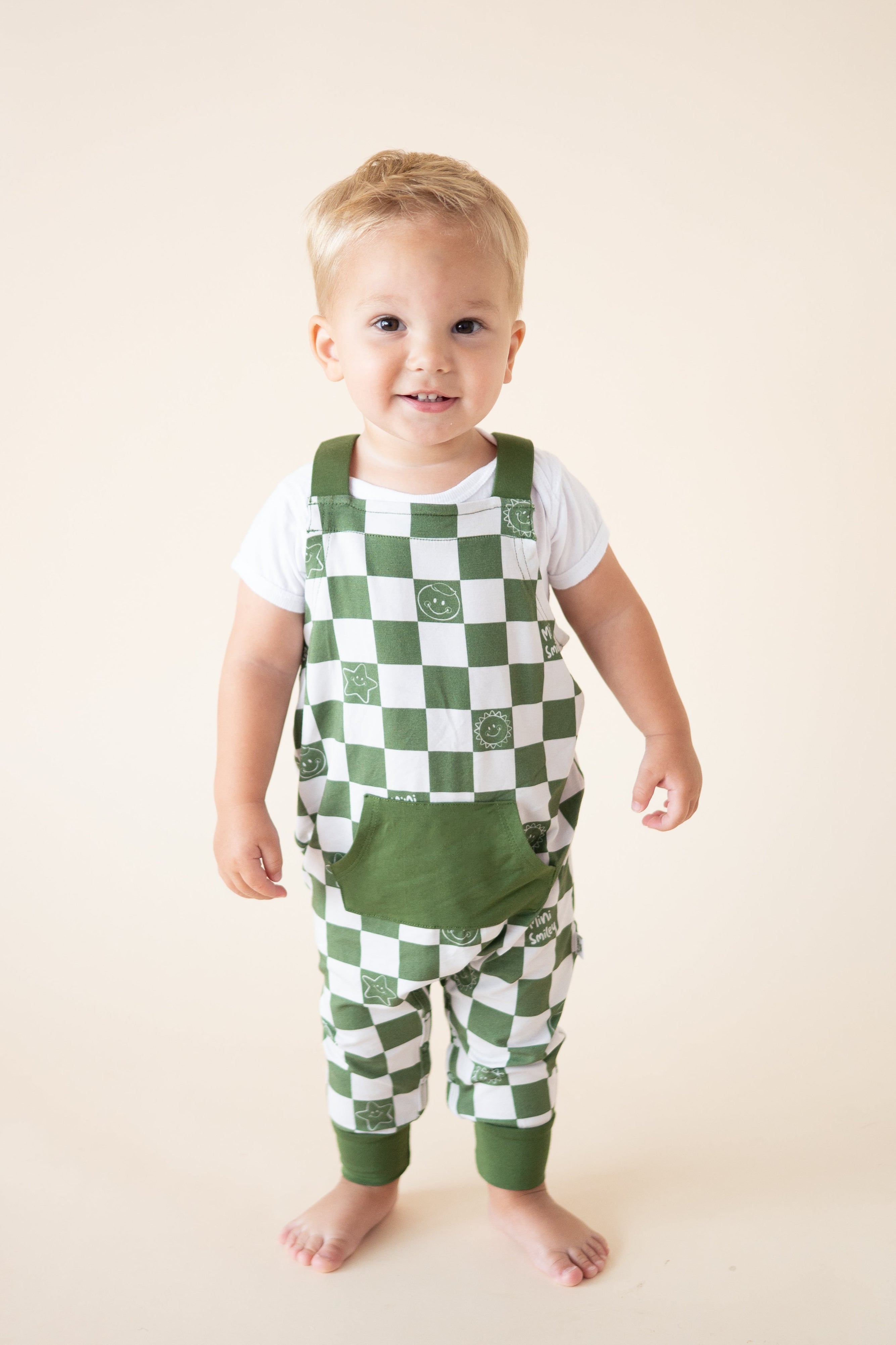 A toddler boy wears green and white checkered overalls with smiley faces over a white t-shirt.