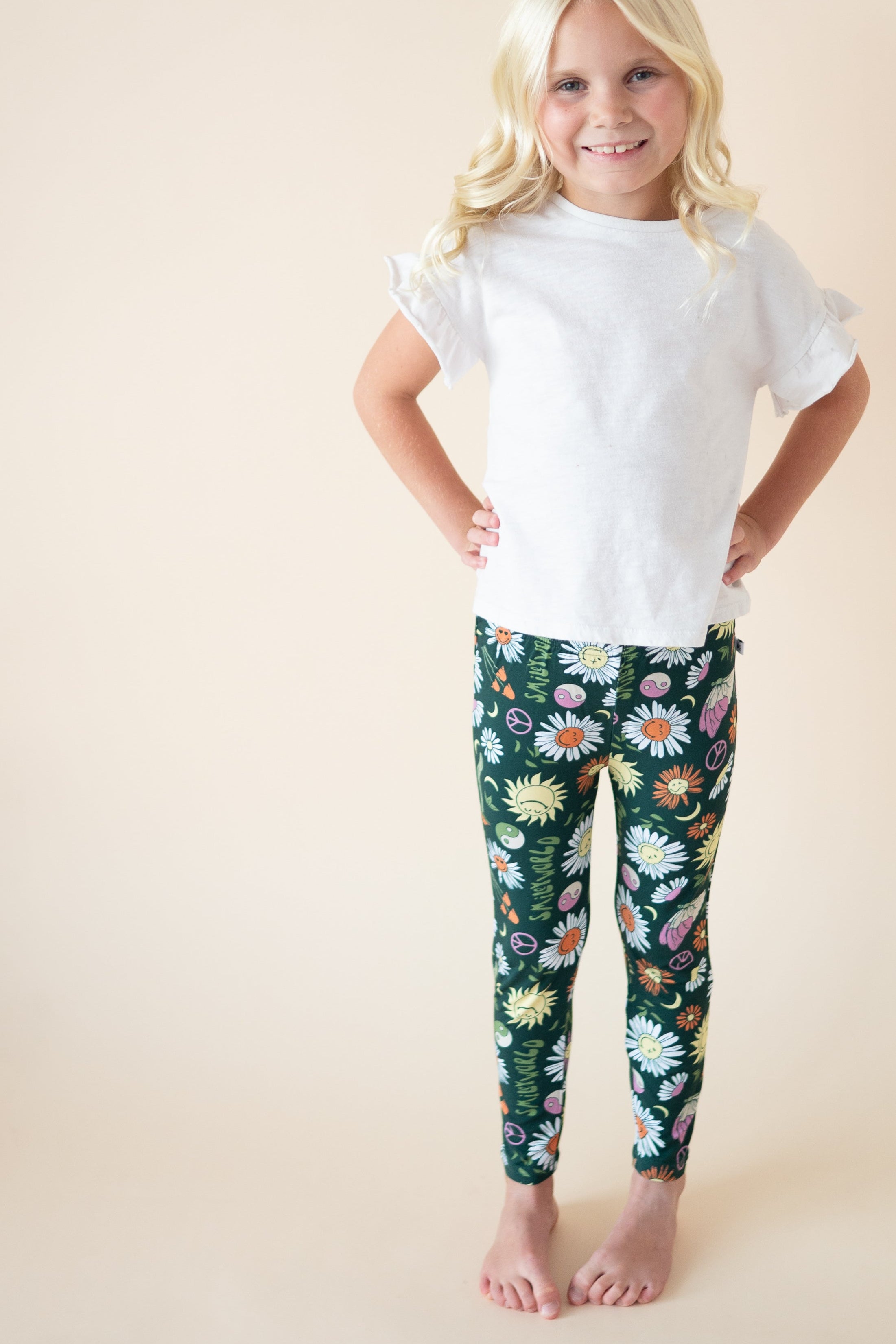 A young girl wears white t-shirt and green leggings with a smiley face and floral print.