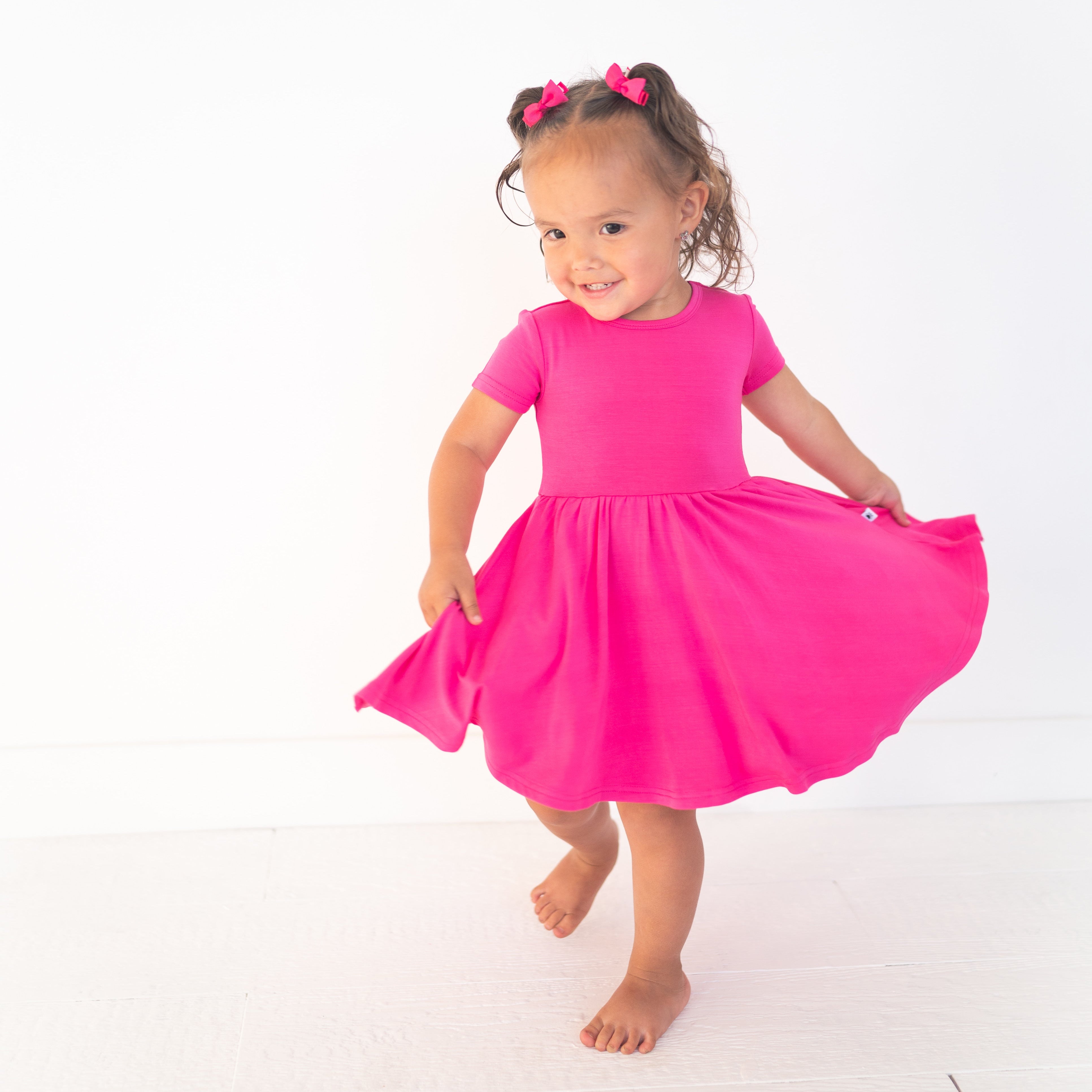 A young girl twirls in a bright pink bamboo dress with two pink bows in her hair.