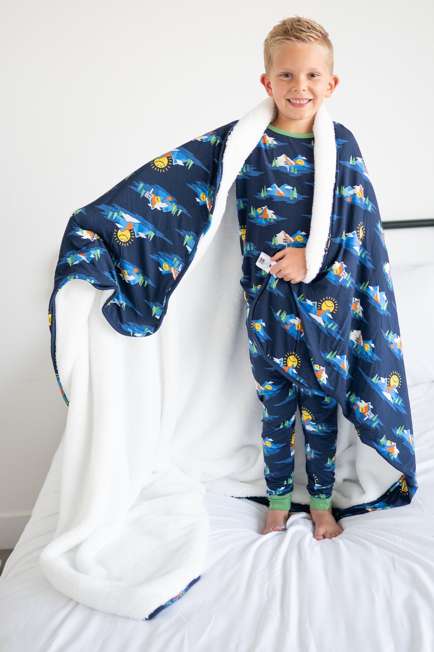 A boy wears a navy blue blanket with a SmileyWorld mountain print and white trim.