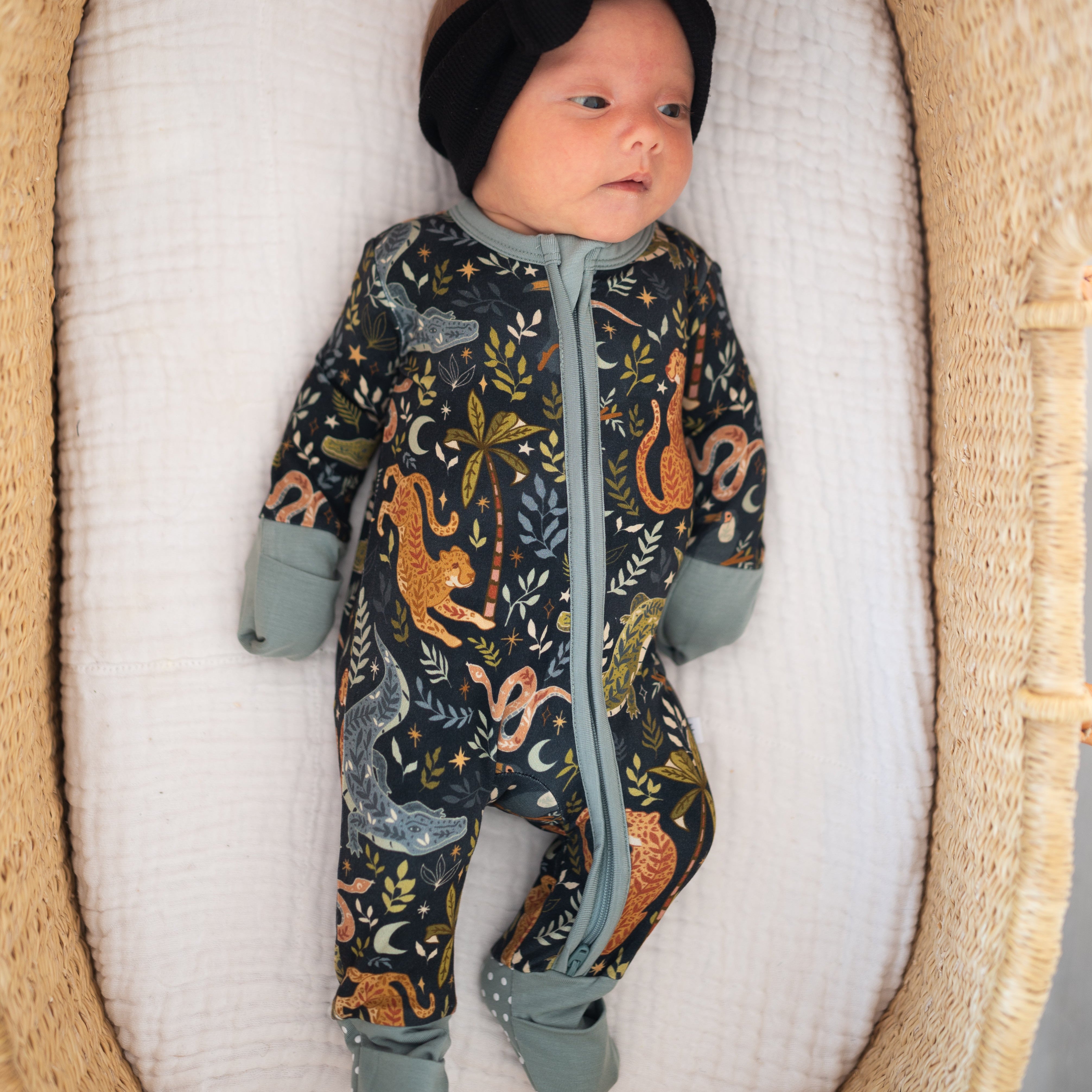 Newborn baby in a patterned onesie and black headband with a bow, lying in a wooden crib.