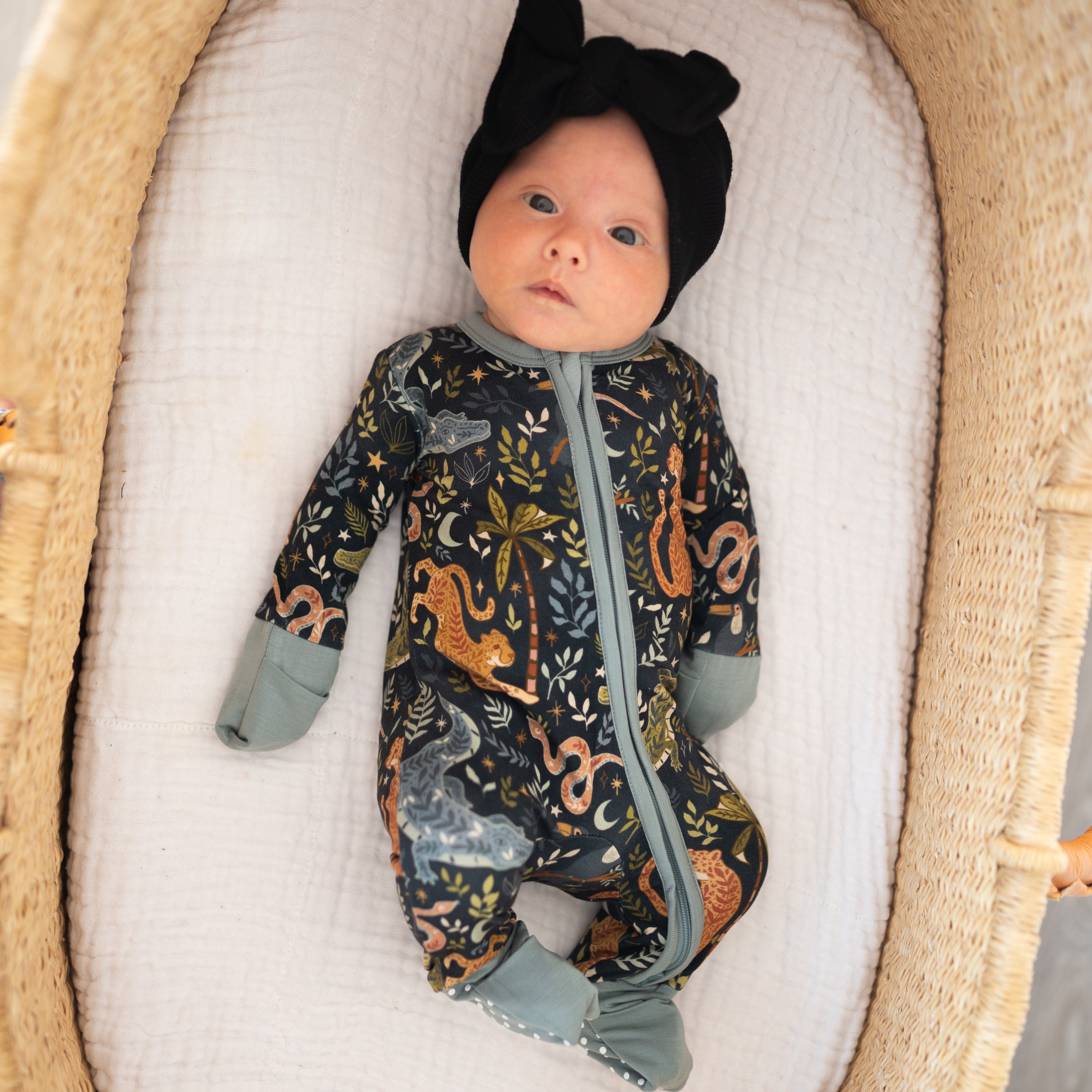 Baby in a patterned onesie and black hat lying in a wooden crib with white mattress.