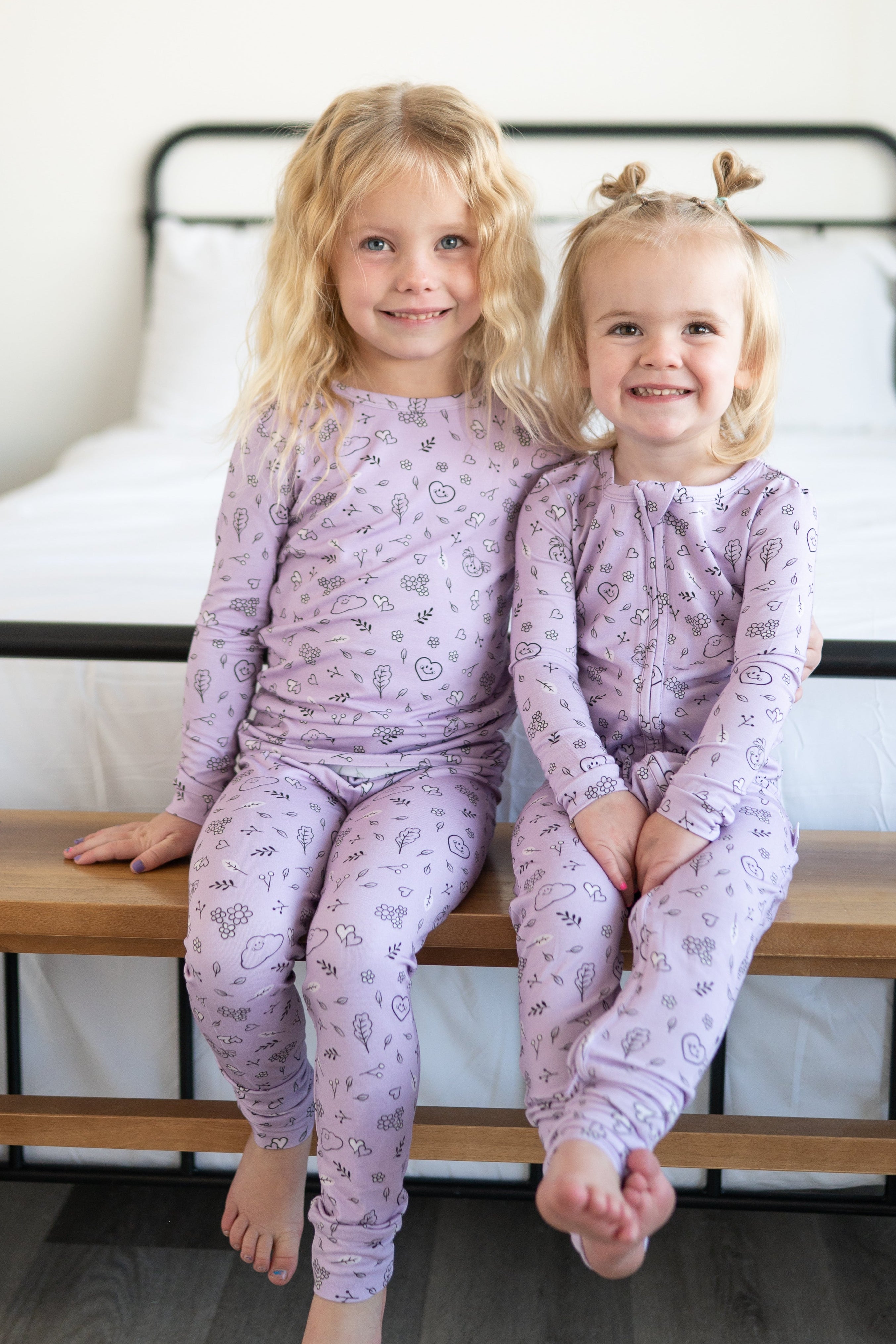 Two young girls in lilac doodle print bamboo pajamas sit on a wooden bench.
