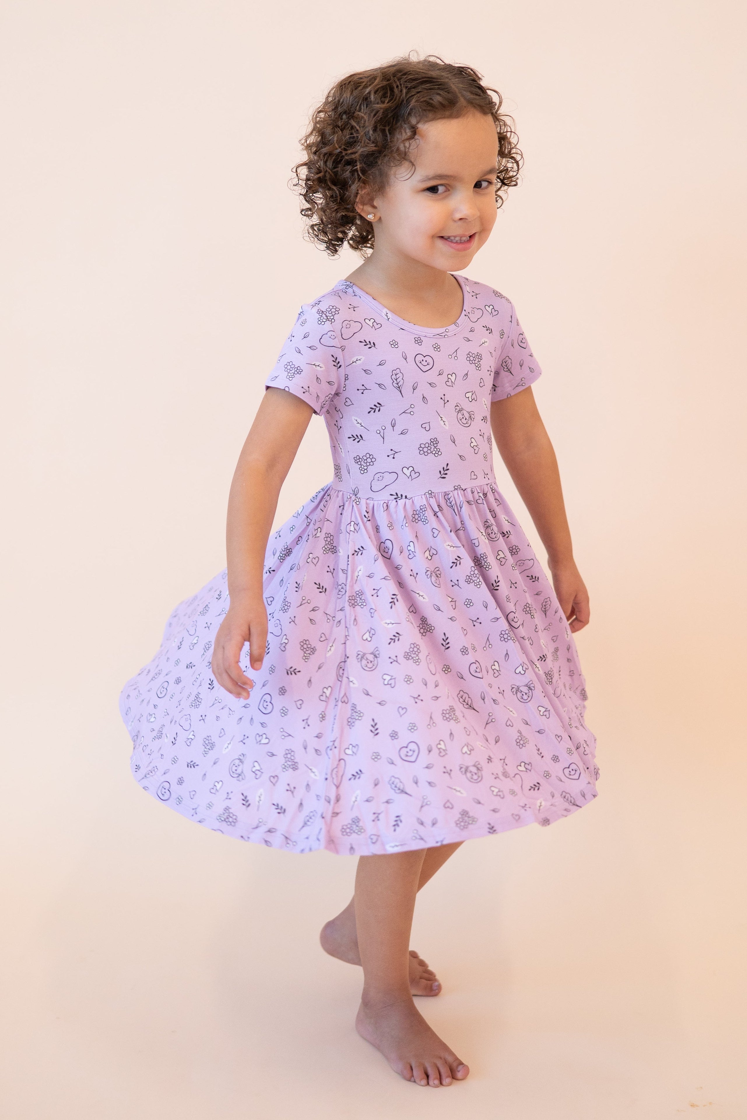 A young girl in a lilac dress with a doodle print smiles and twirls.