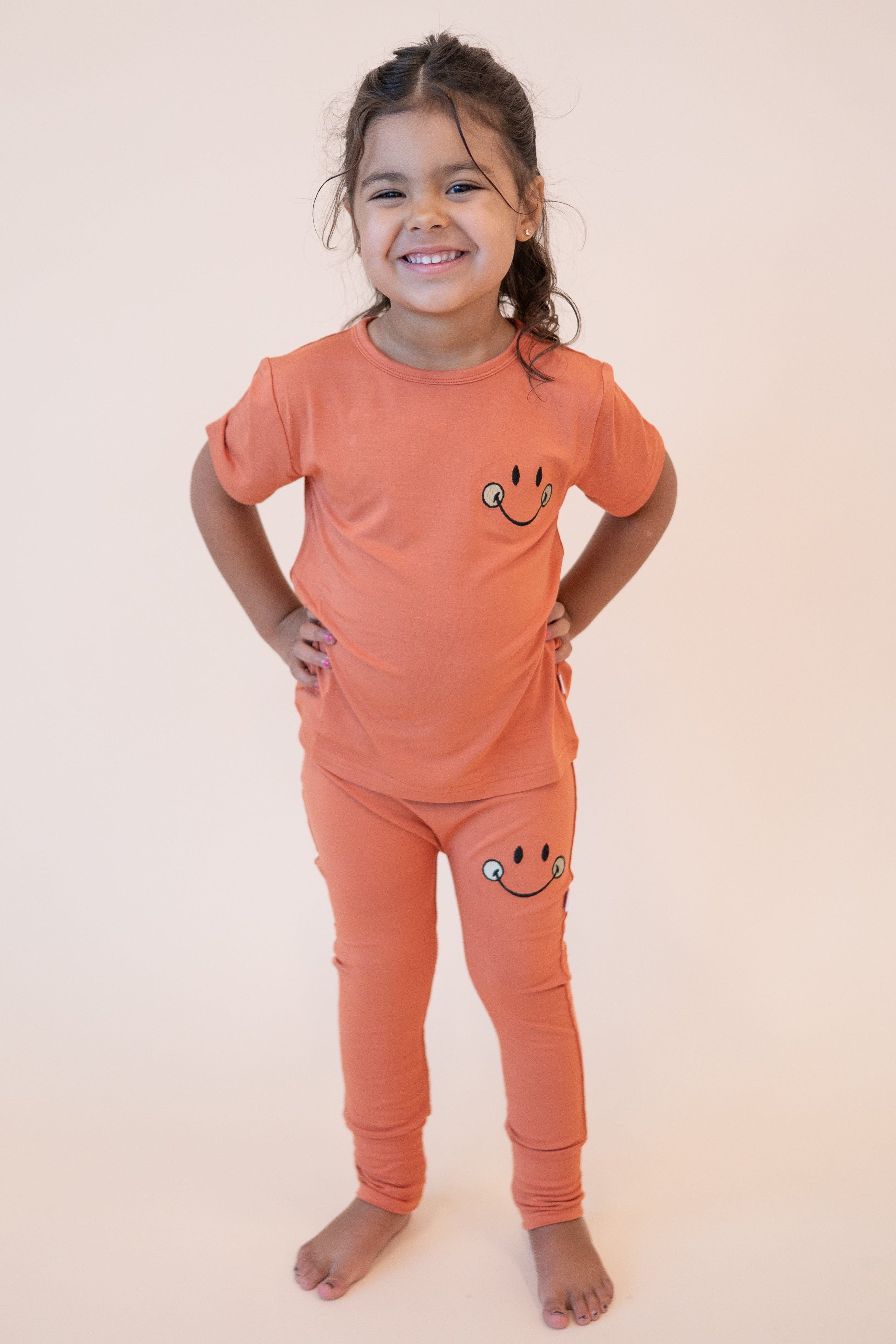 A young girl wears orange bamboo pants and a matching t-shirt with a smiley face design.