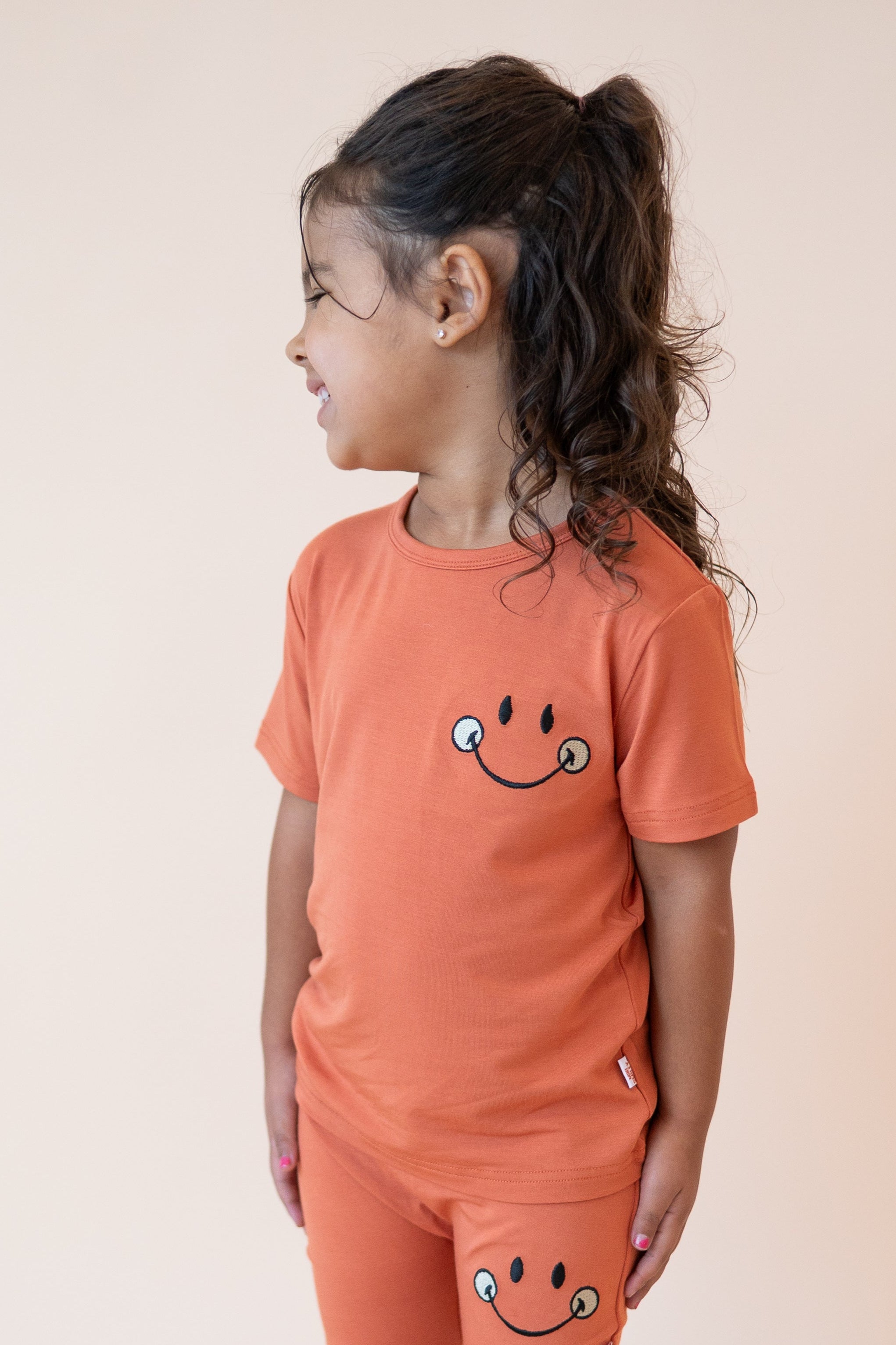 A young girl wears an orange t-shirt with a smiley face graphic.