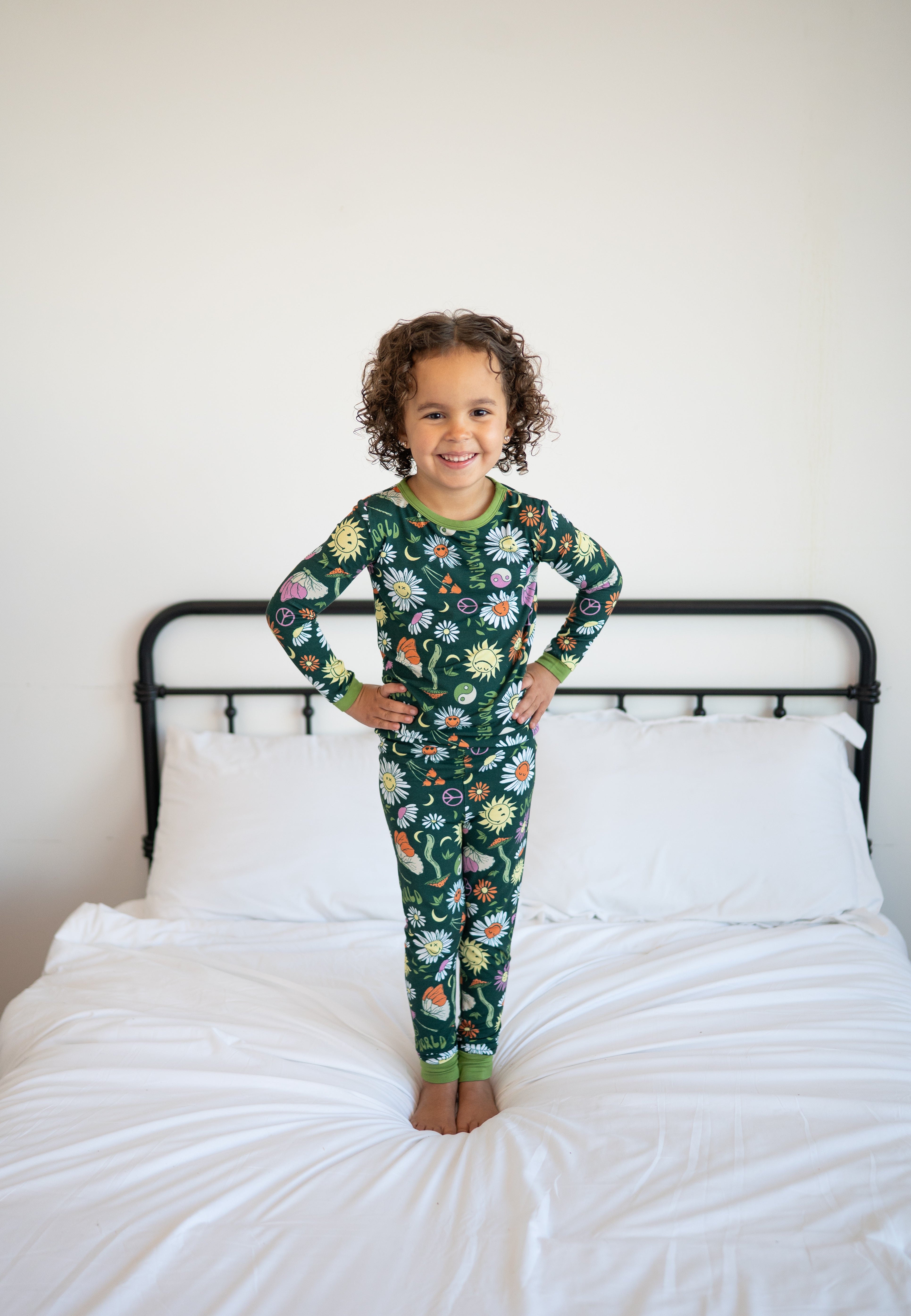A young child stands on a bed wearing green pajamas with a floral and smiley face print.
