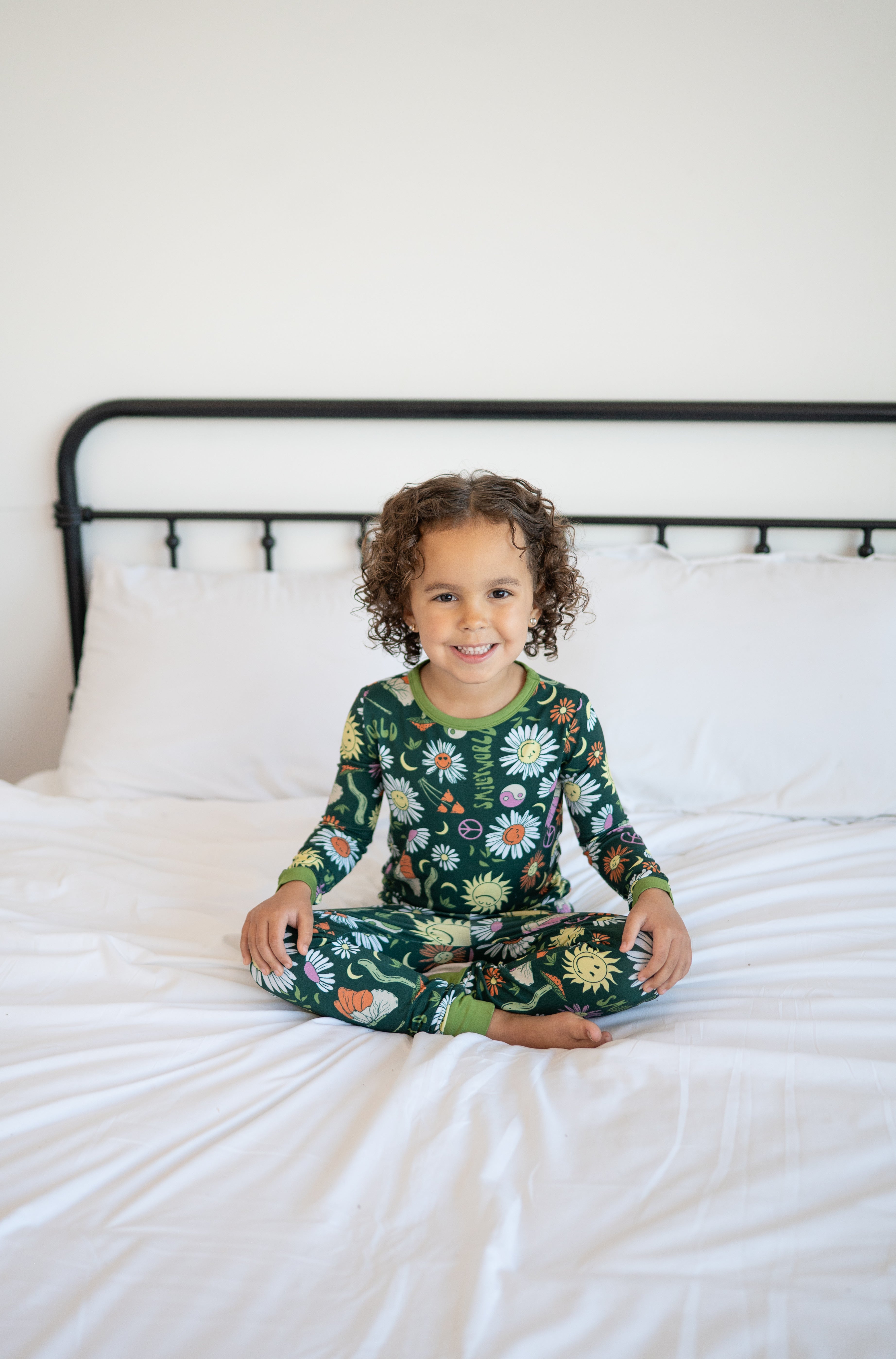 A young child sits cross-legged on a bed wearing green pajamas with a floral and smiley face print.
