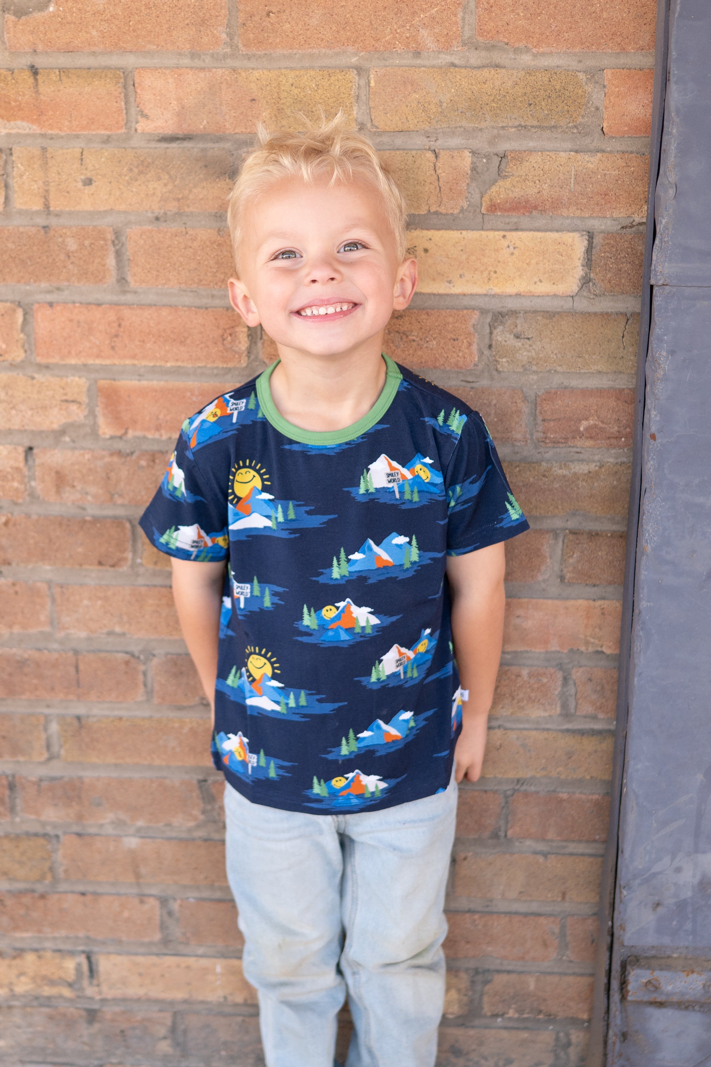 A young boy smiles in a navy t-shirt with a SmileyWorld mountain print and light blue jeans.