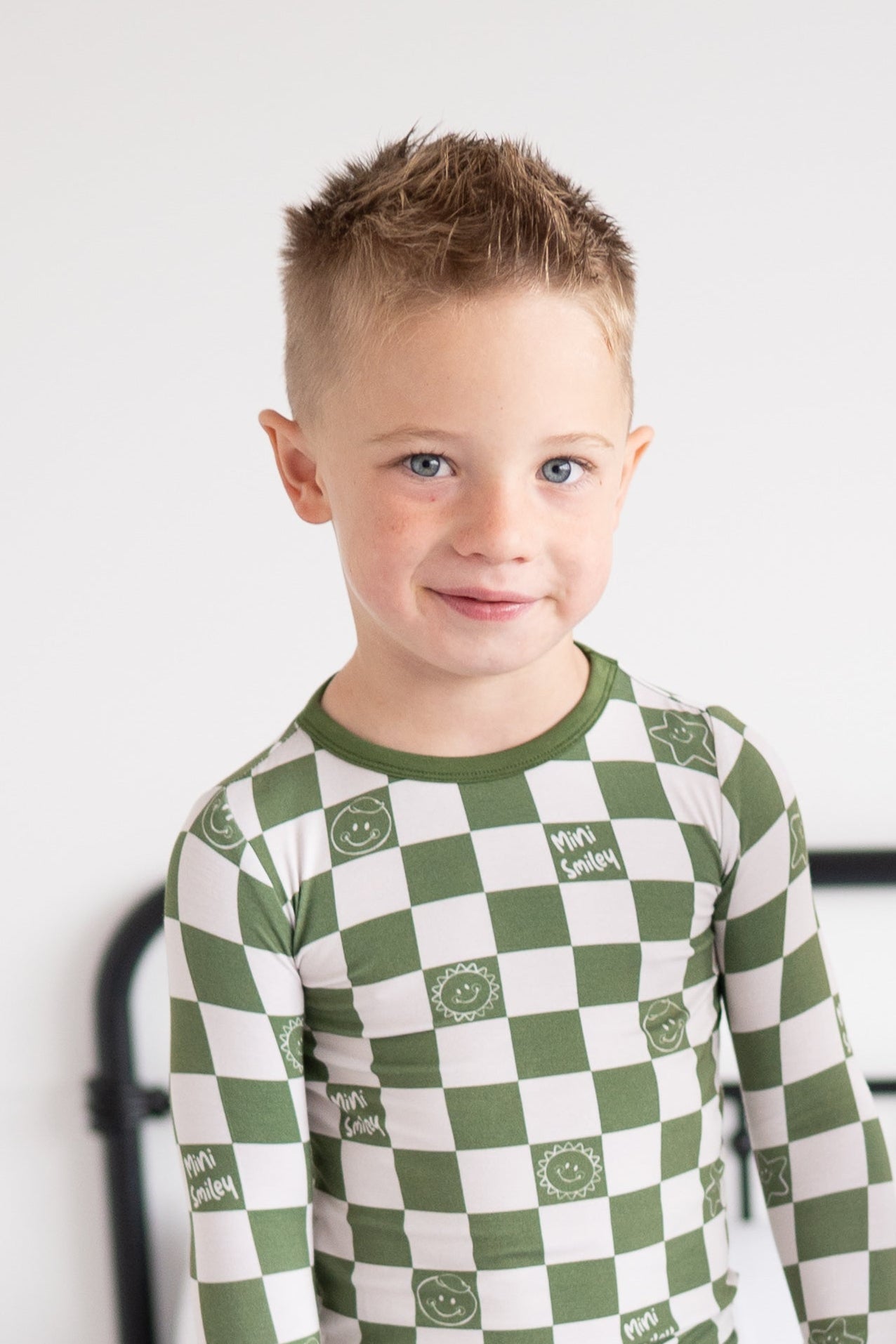 A young boy smiles wearing a green and white checkered long-sleeved pajama set with smiley face prints.