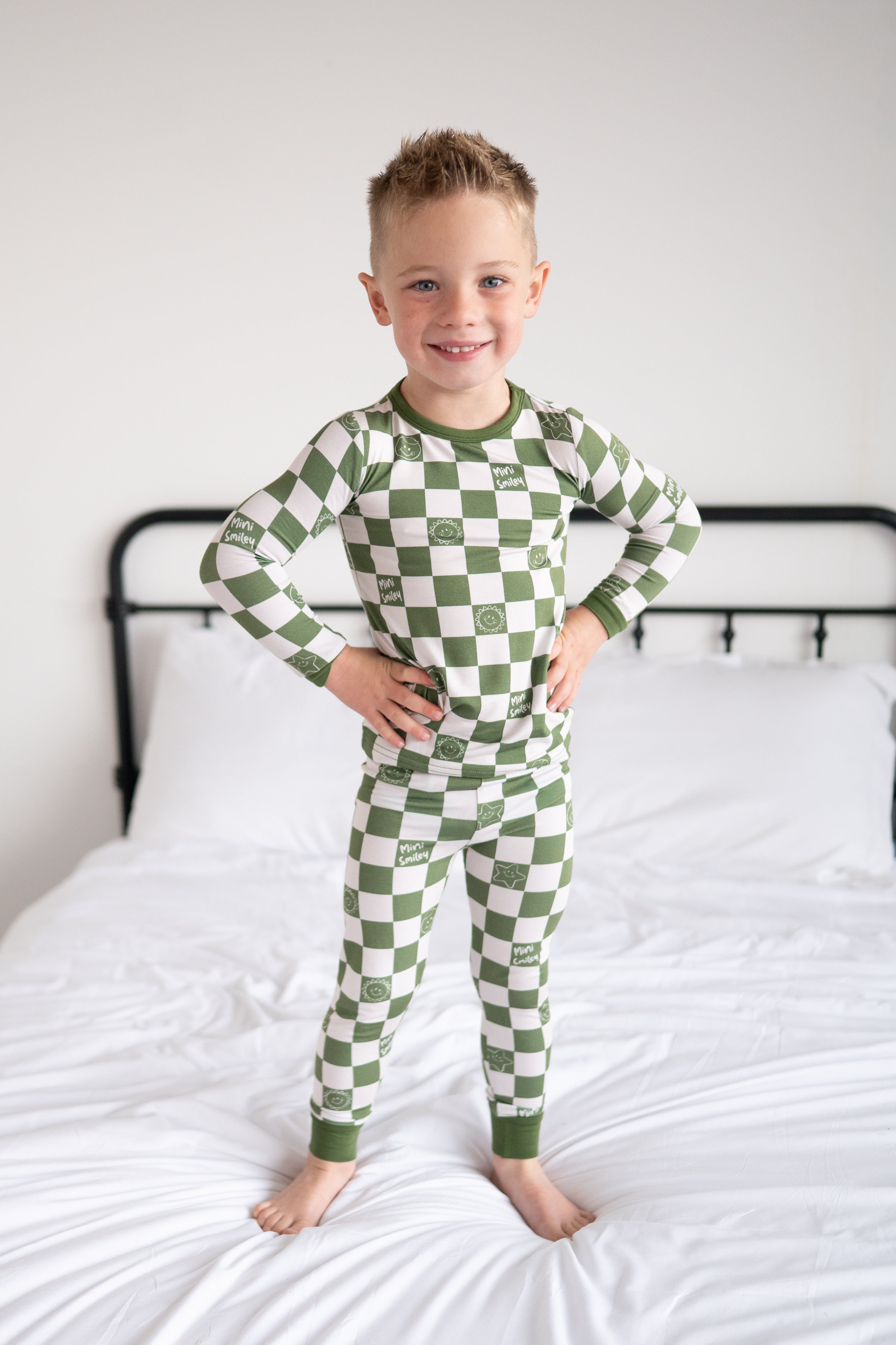 A young boy stands on a bed wearing green and white checkered pajamas with smiley face prints.
