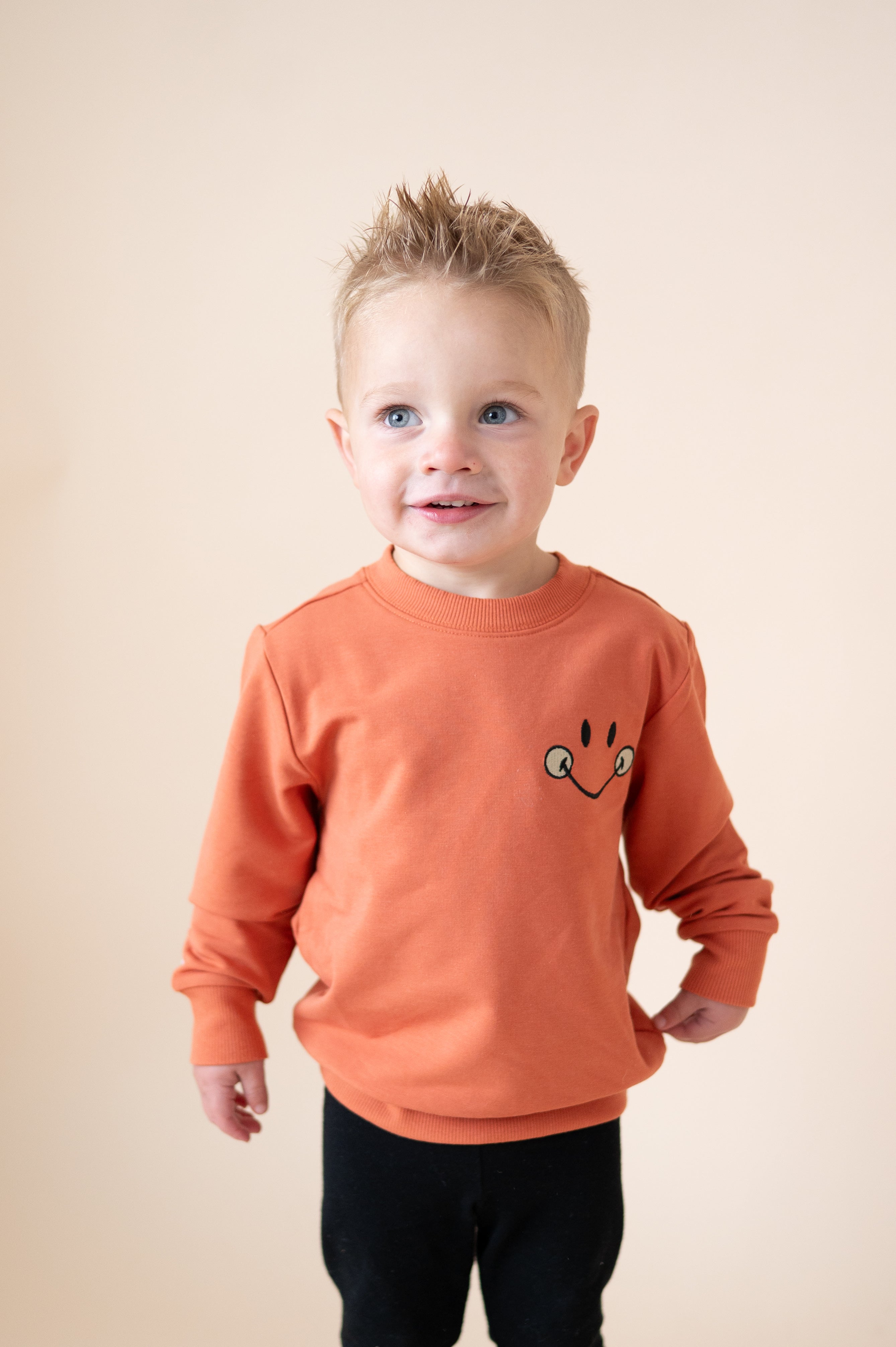 A young boy wears an orange crewneck sweatshirt with a smiley face graphic.