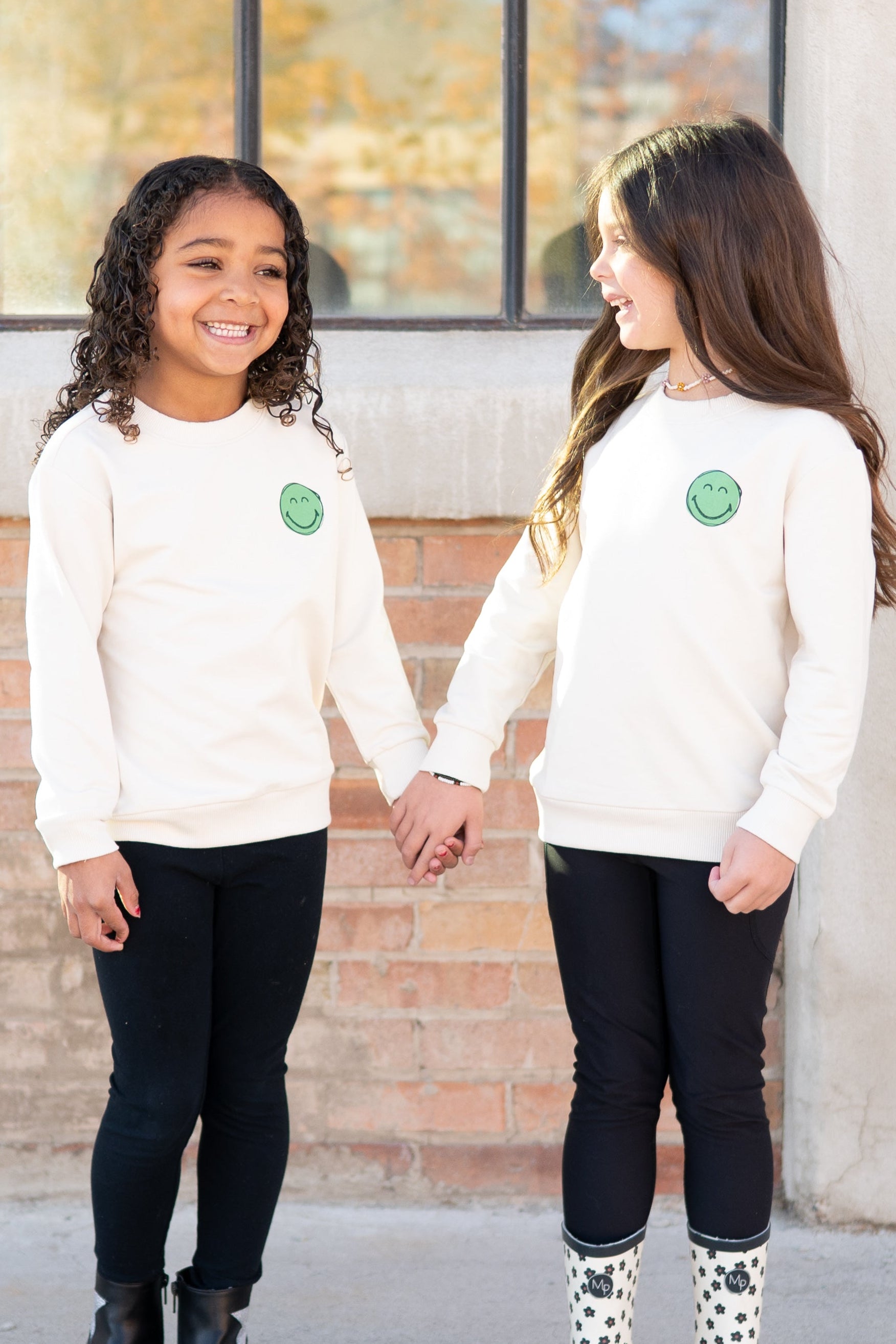 Two girls in cream smiley face sweatshirts and black leggings hold hands.