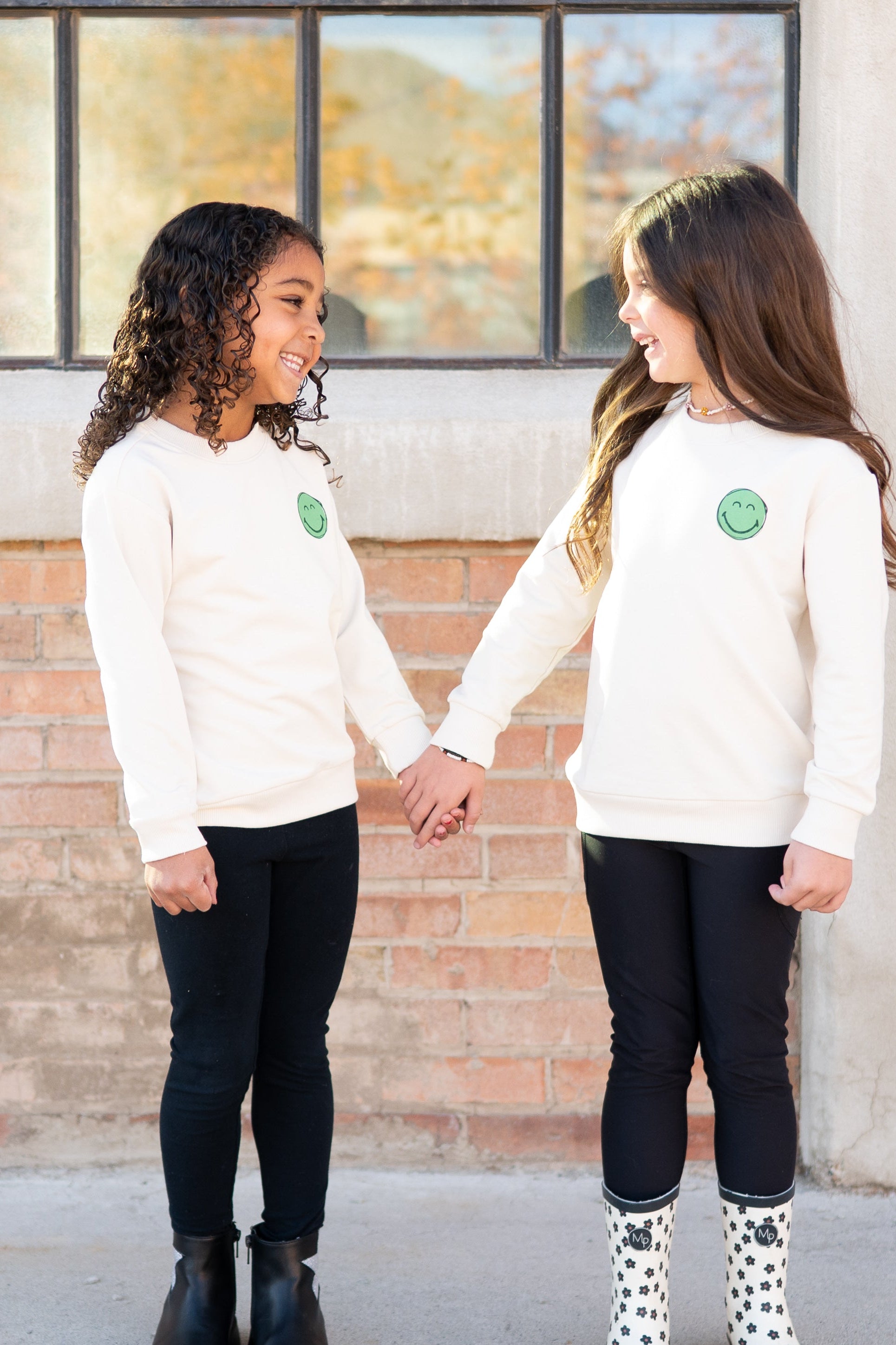 Two girls in cream smiley face sweatshirts and black leggings hold hands.