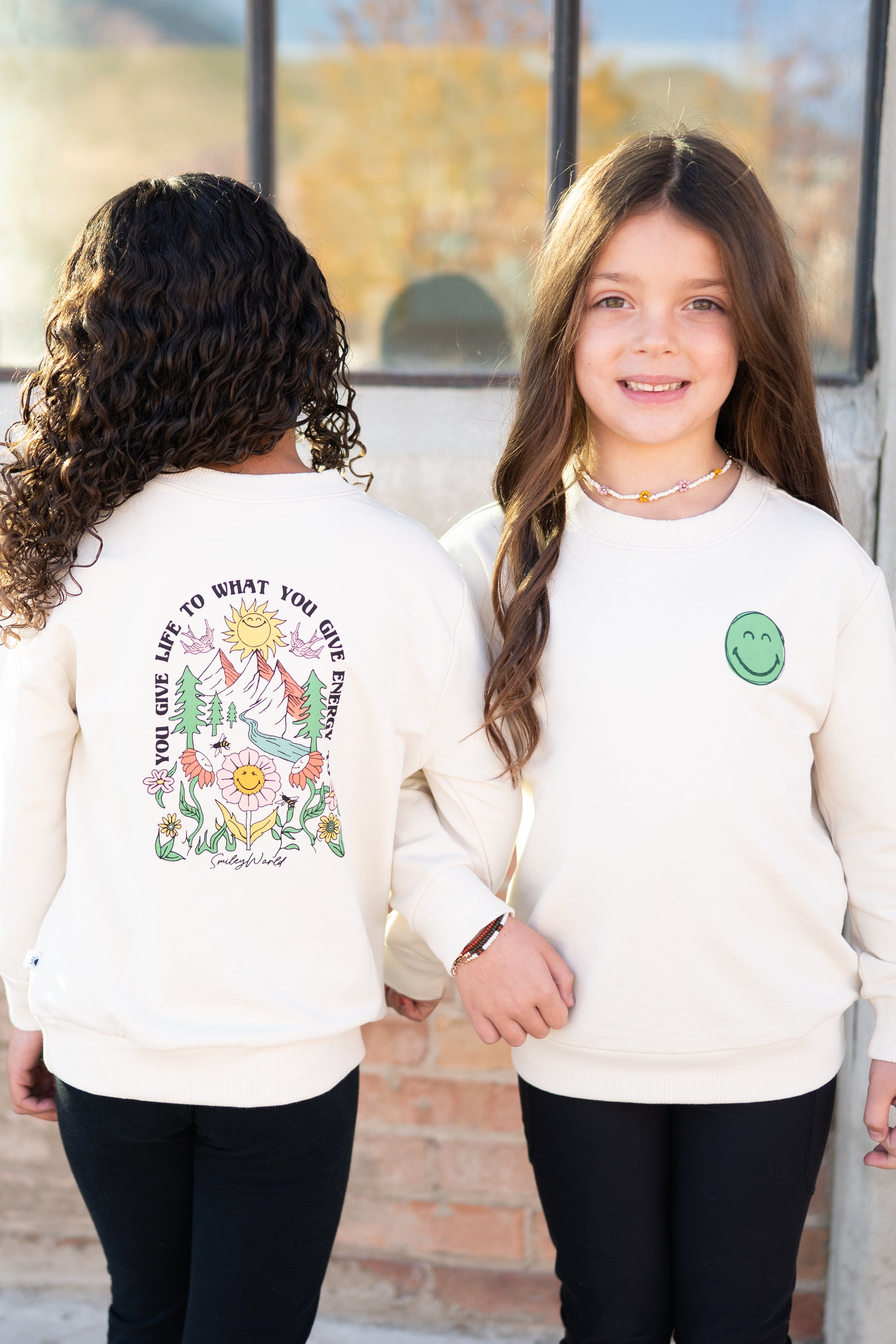 Two girls wear cream-colored crewneck sweatshirts with SmileyWorld graphics.