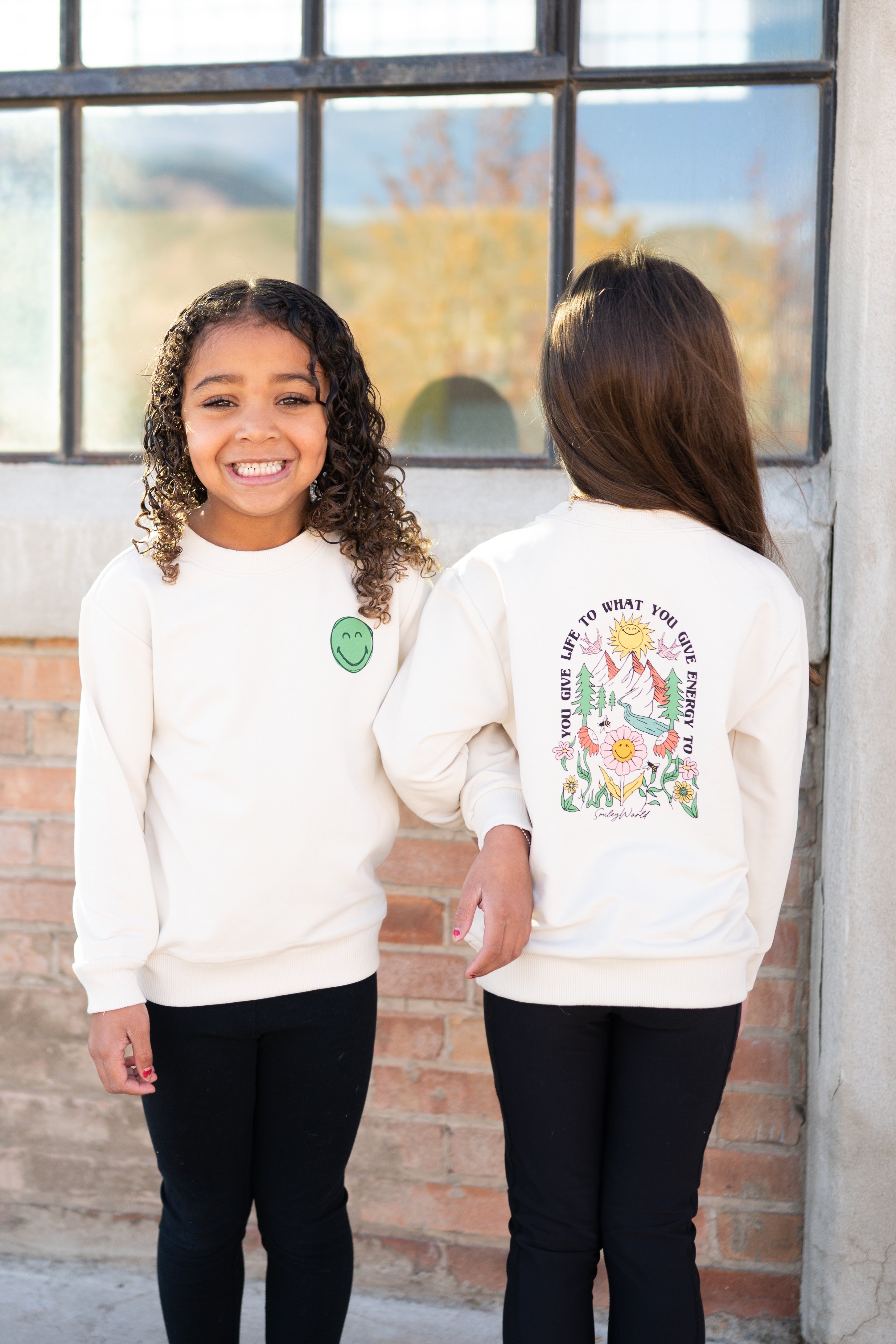 Two girls wear cream crewneck sweatshirts with SmileyWorld graphics.