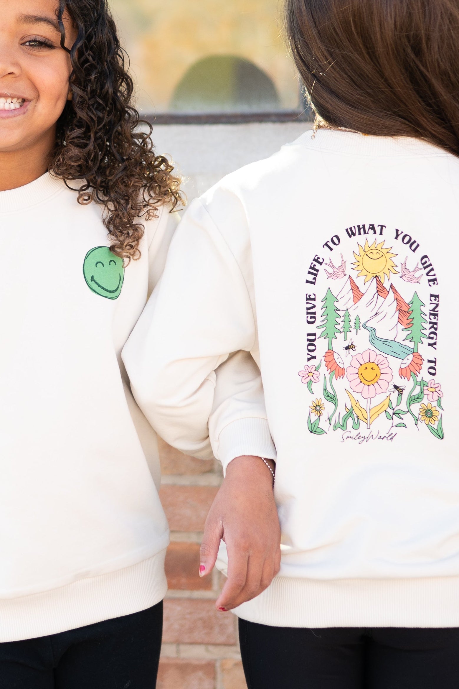 Cream crewneck sweatshirt with a green smiley face on the left chest and a colorful nature graphic on the back.