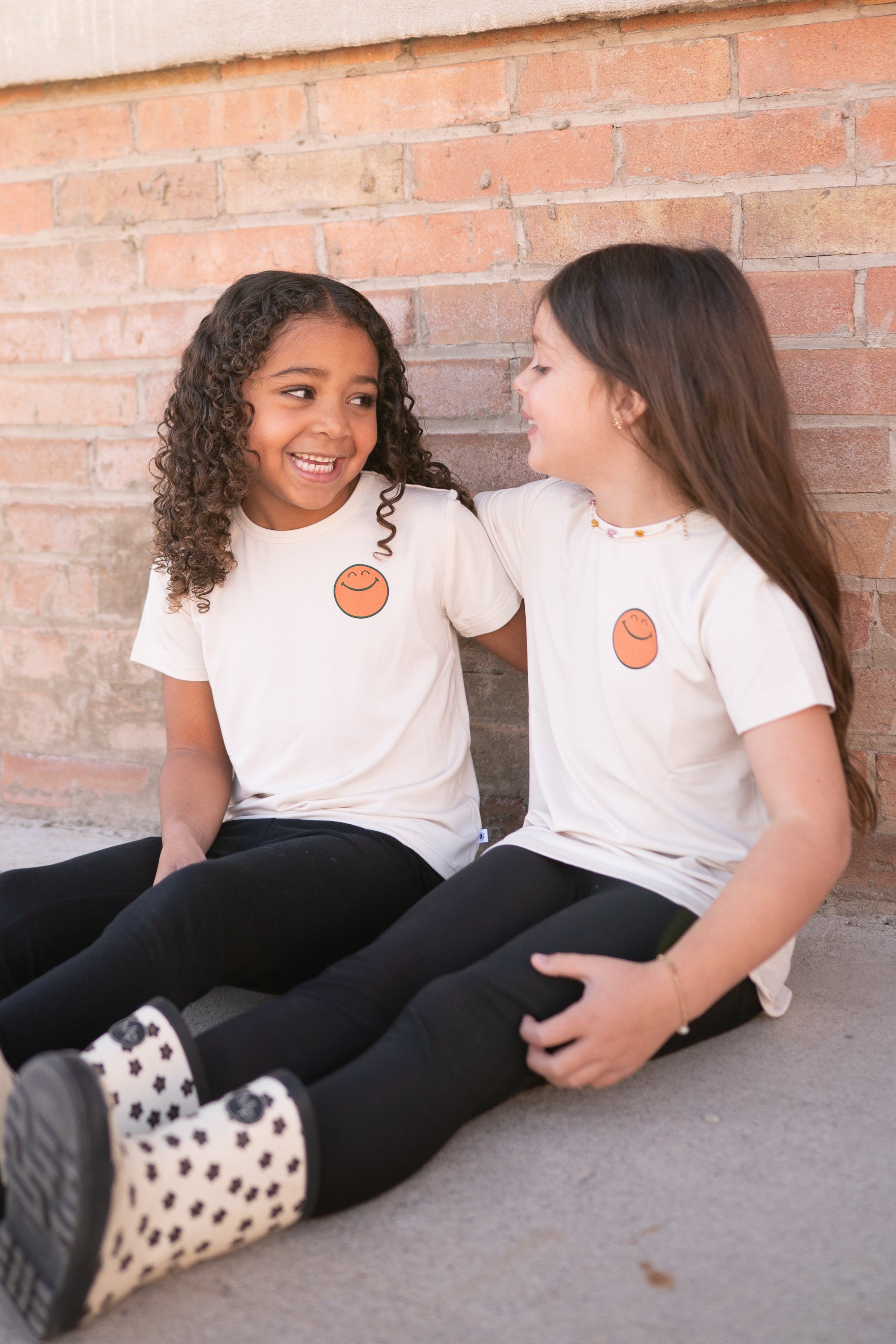 Two girls in cream t-shirts with orange smiley faces sit against a brick wall.