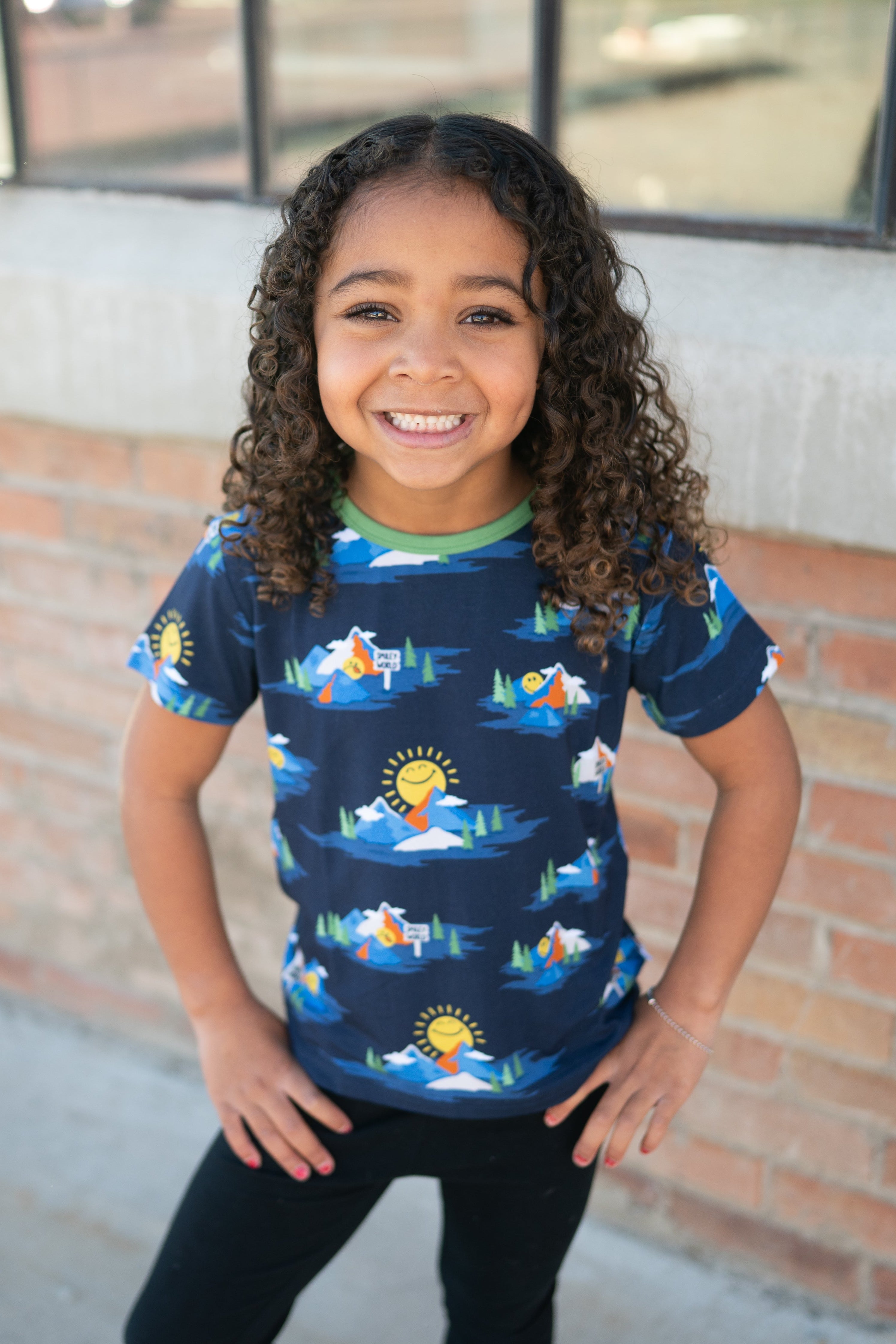 A young girl smiles wearing a navy t-shirt with a SmileyWorld mountain print.