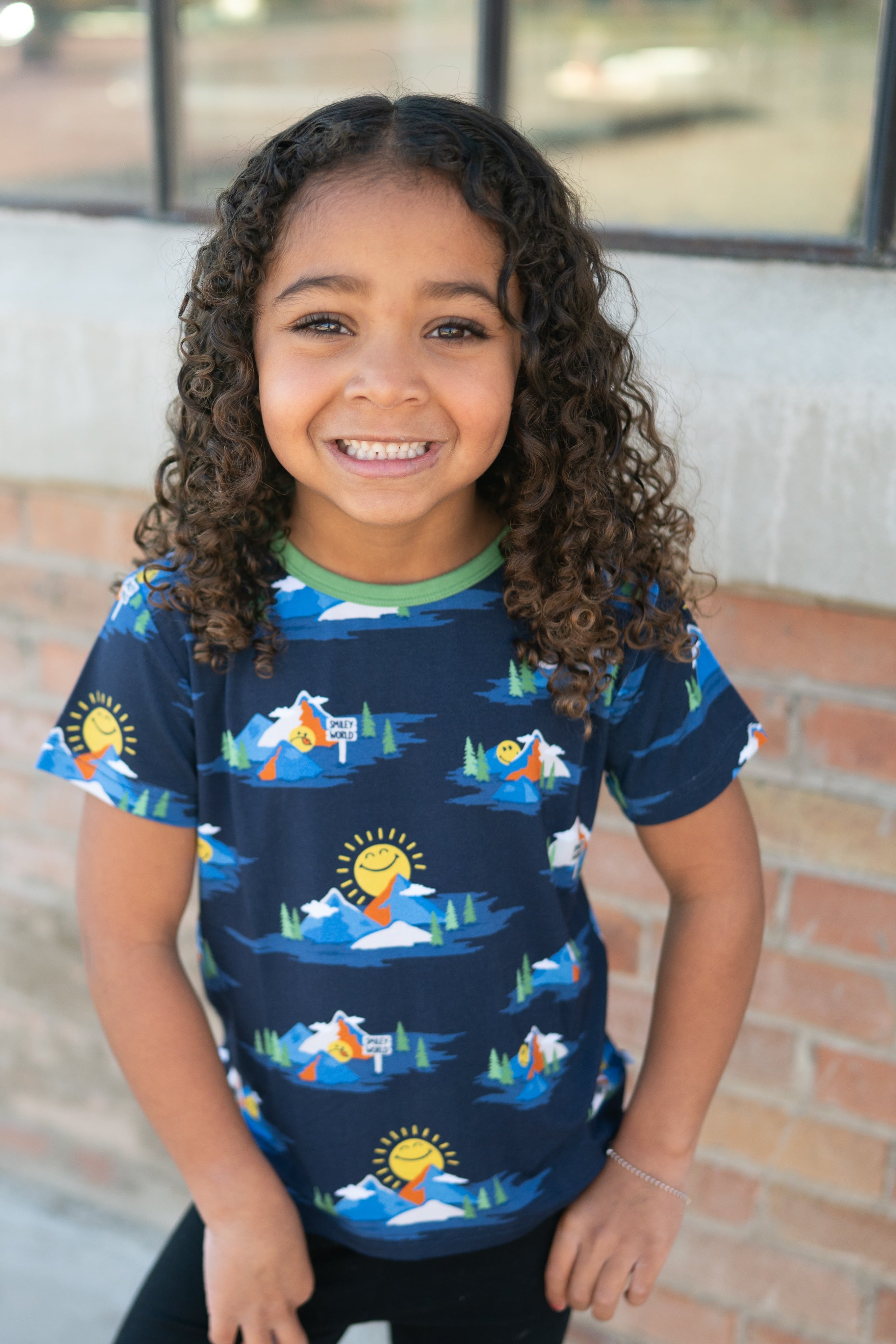 A young girl smiles wearing a navy t-shirt with a SmileyWorld mountain print.