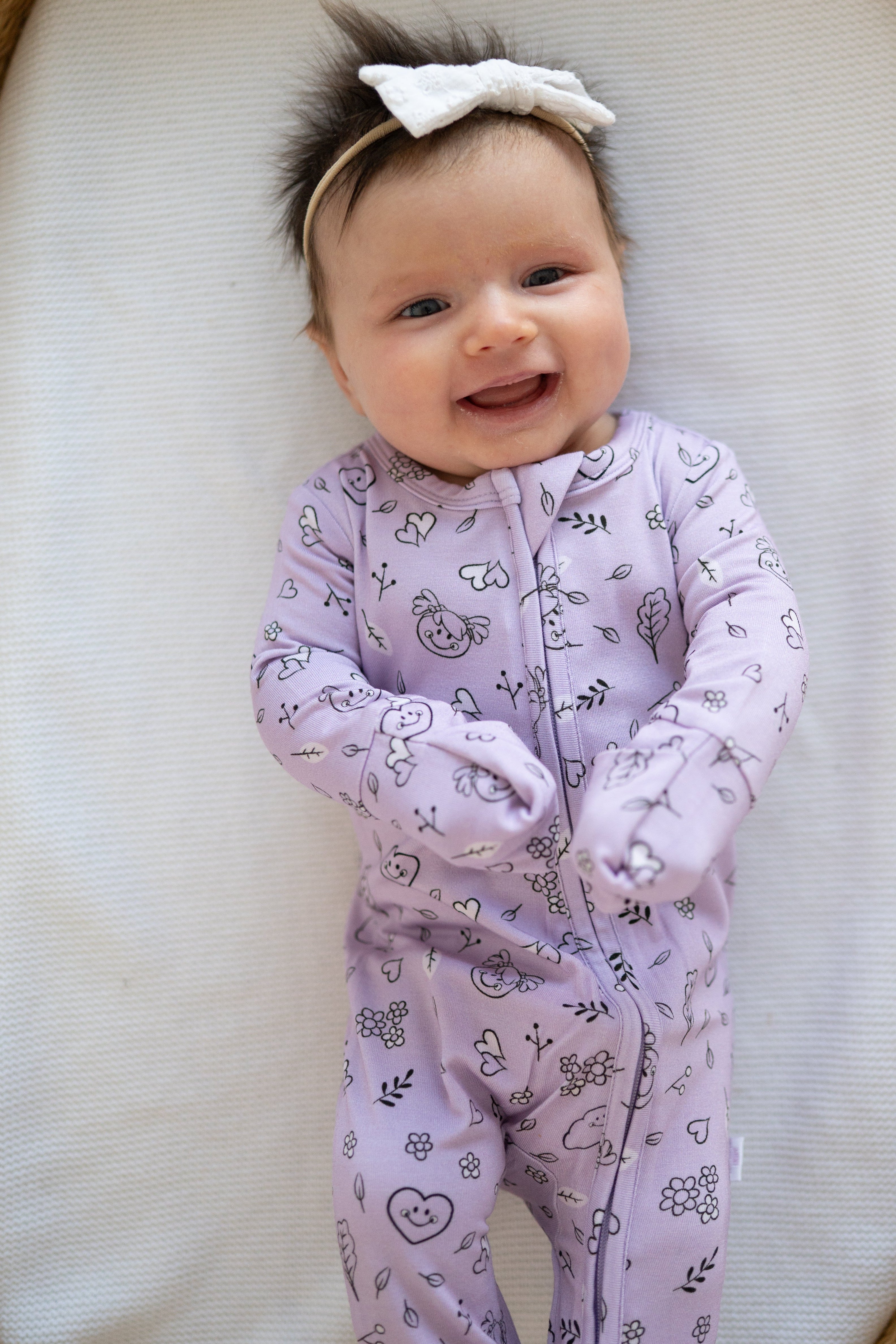 A baby wearing a lilac bamboo sleeper with a doodle print and a white bow.