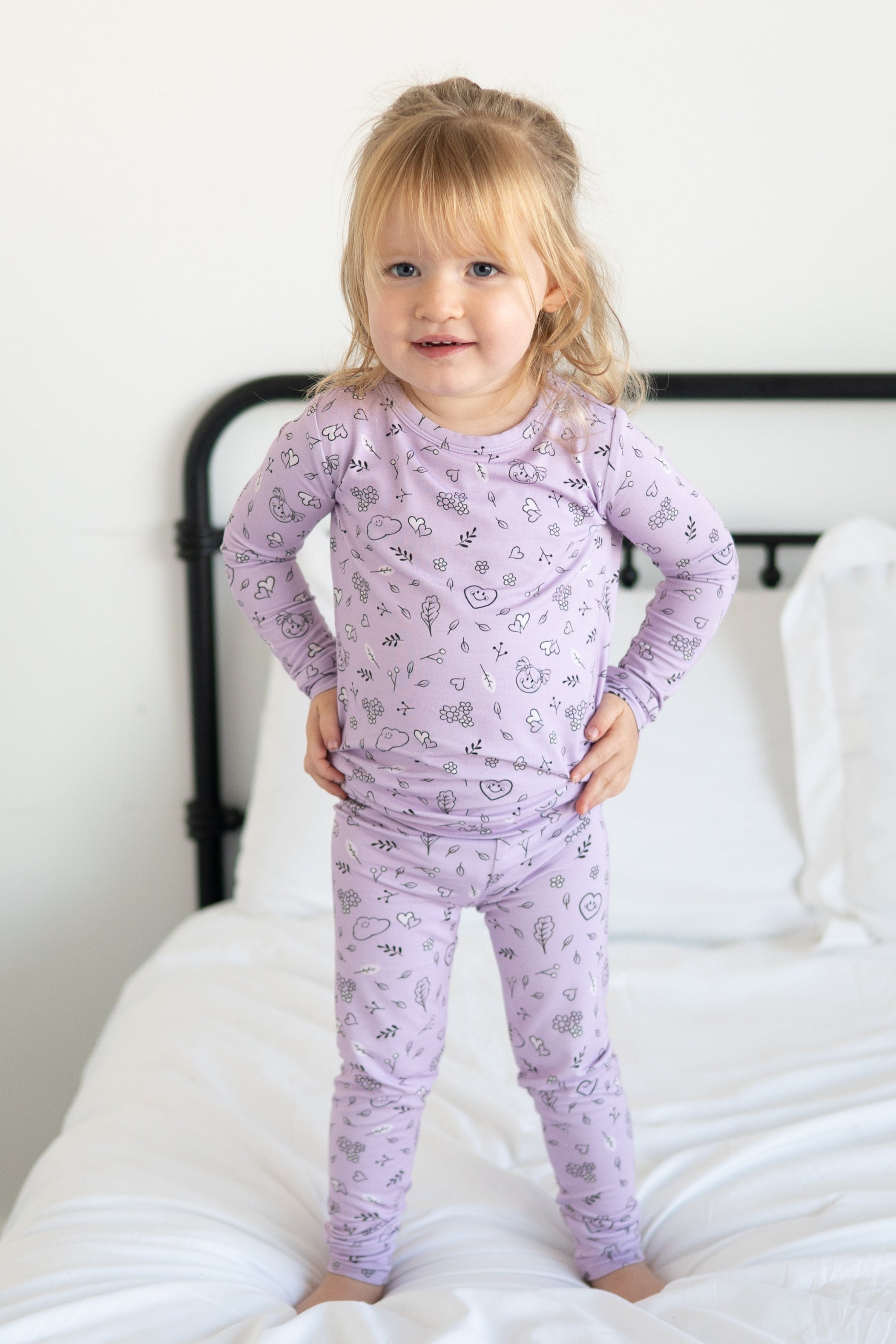 A young girl stands on a bed wearing a lilac pajama set with a doodle print.