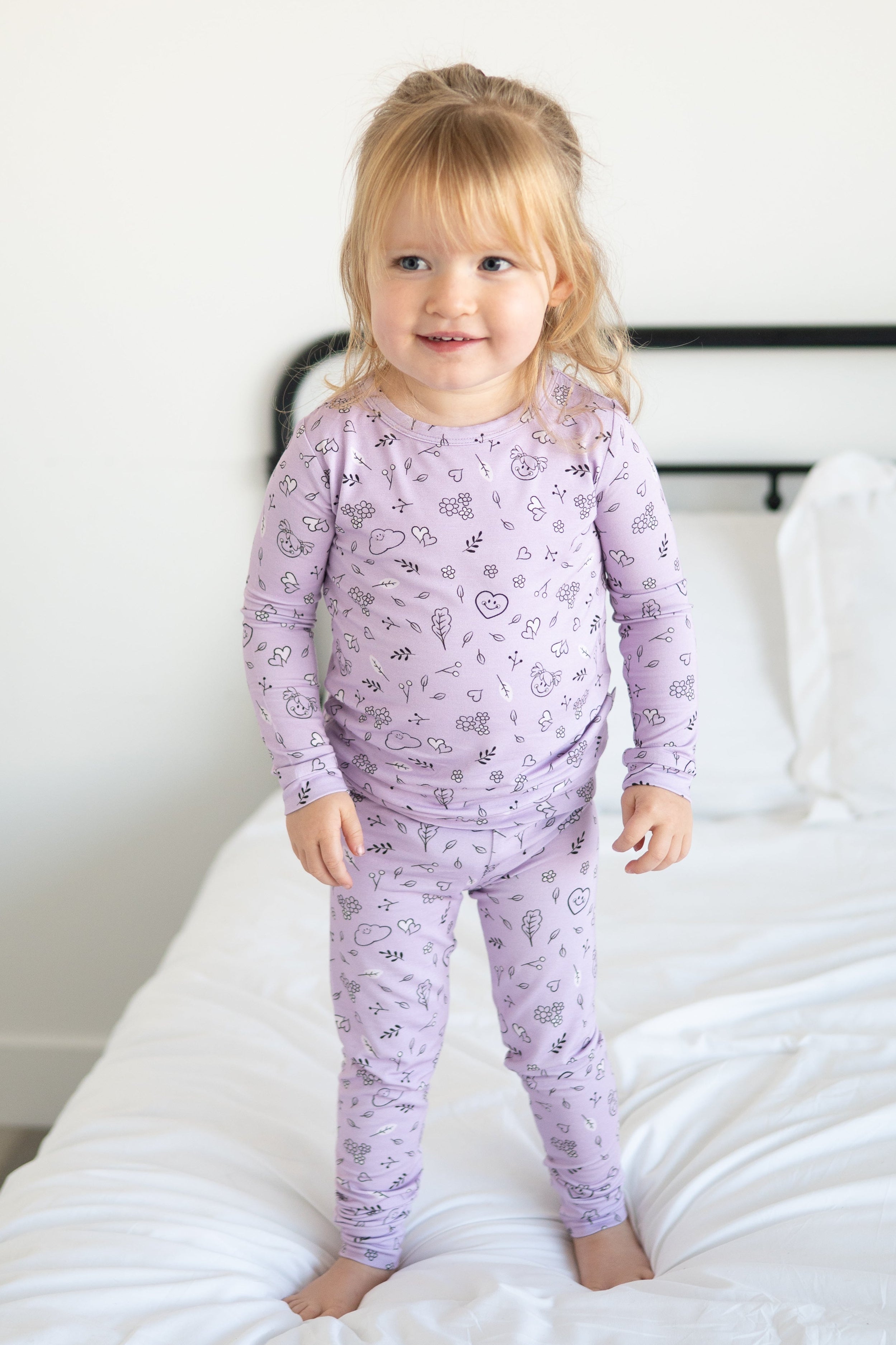 A young girl stands on a bed wearing lilac pajamas with a doodle print.