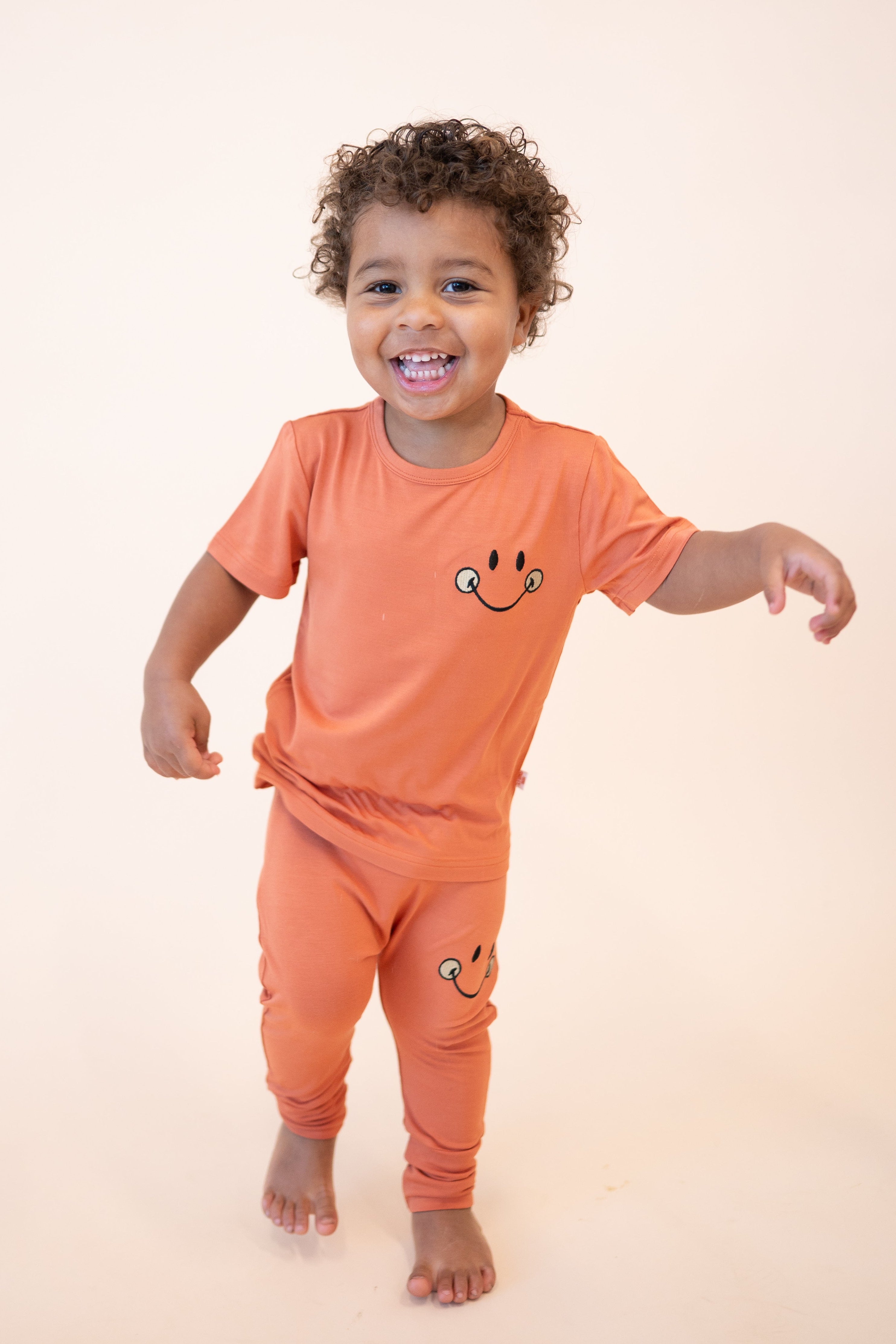 A young child wears an orange t-shirt and pants with a smiley face design.