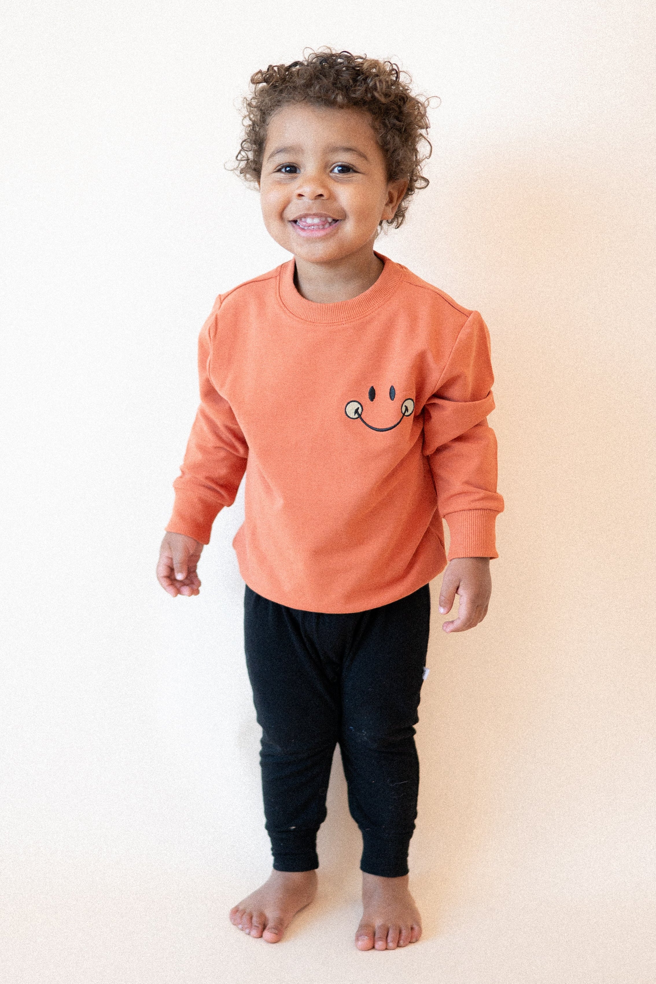 A child wears a rust-colored crewneck sweatshirt with a mini smiley face embroidered on the chest and black pants.