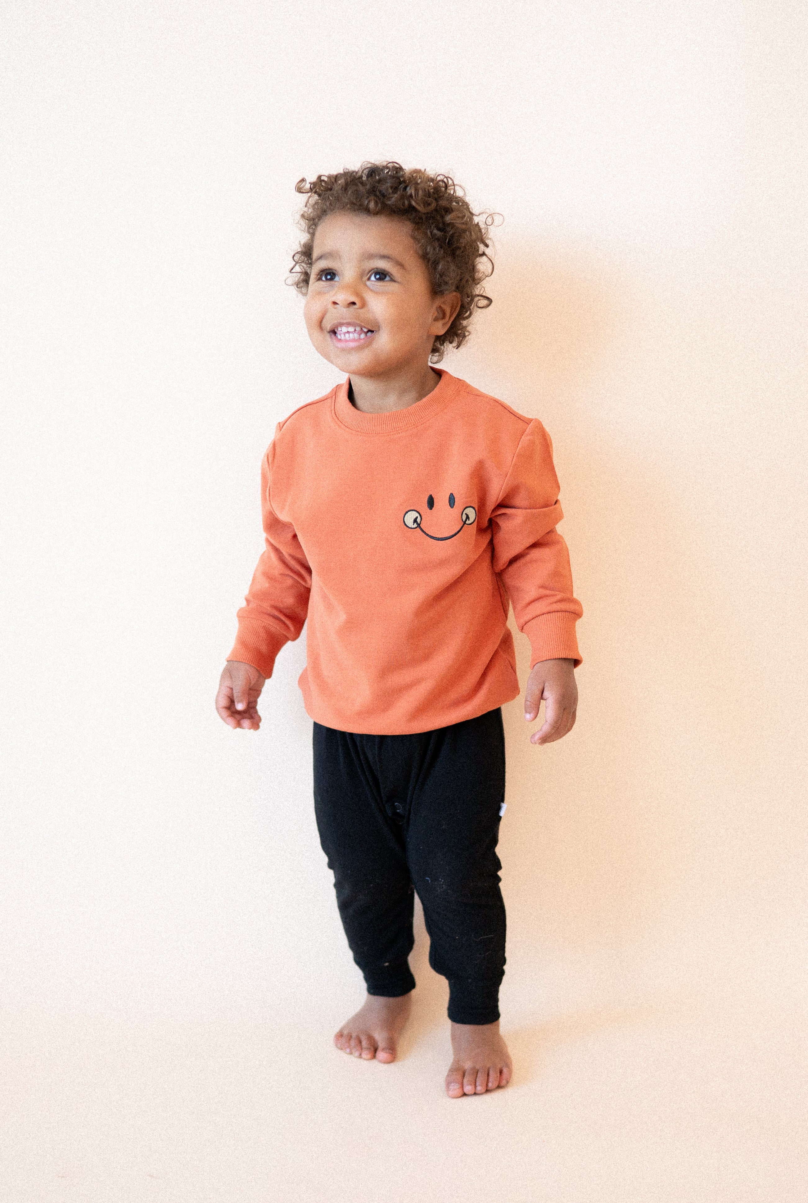 A child wears a rust-colored crewneck sweatshirt with a mini smiley face and black pants.