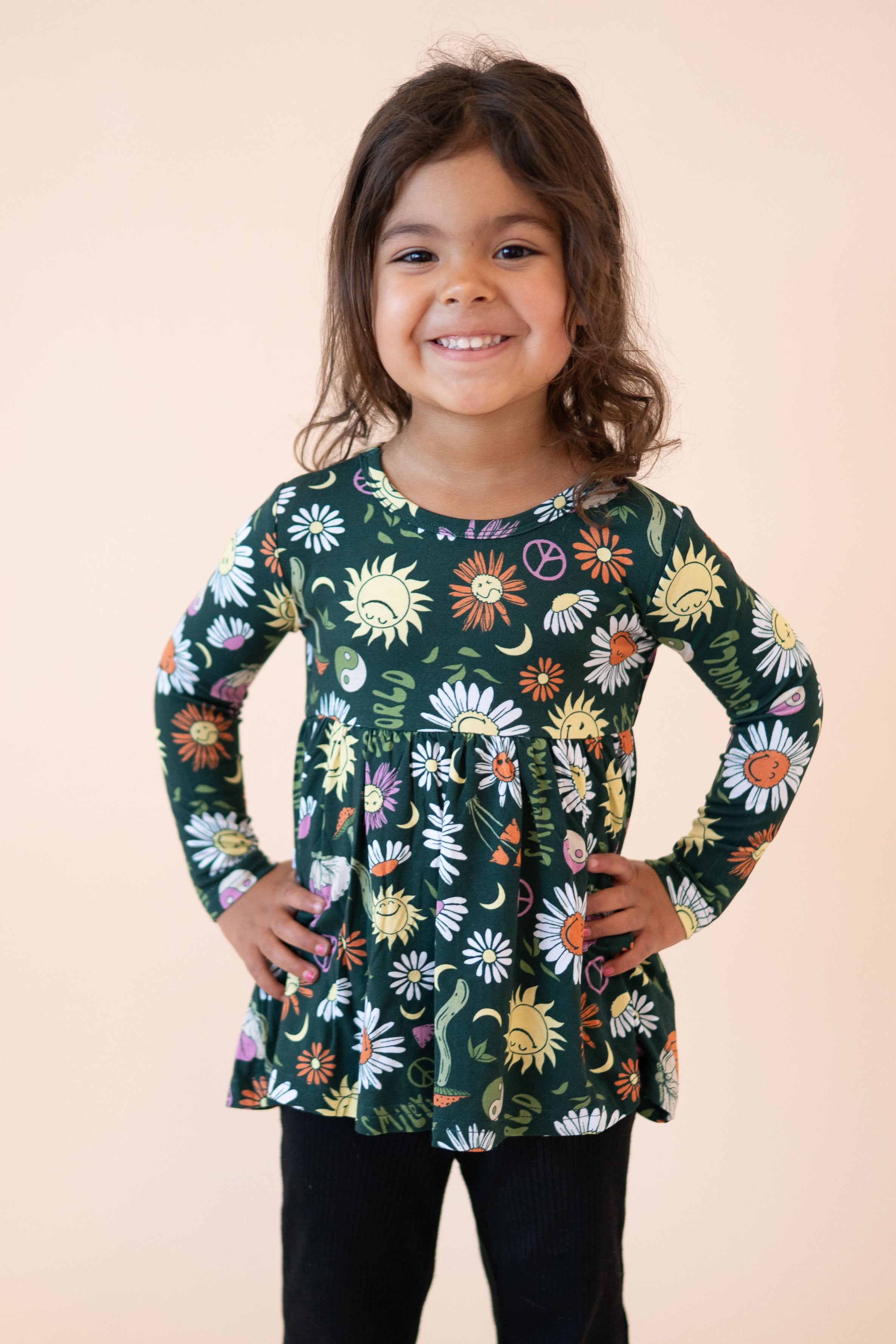 A young girl wears a long-sleeved green peplum top with a groovy floral and smiley face print.