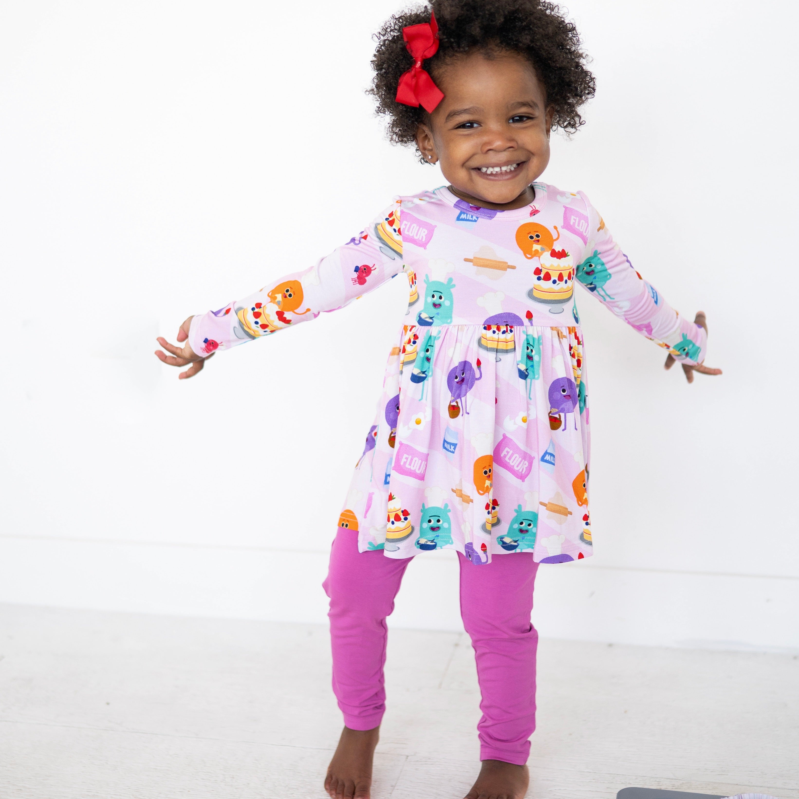 A young girl wears pink leggings and a pink long-sleeved dress with a baking-themed print.