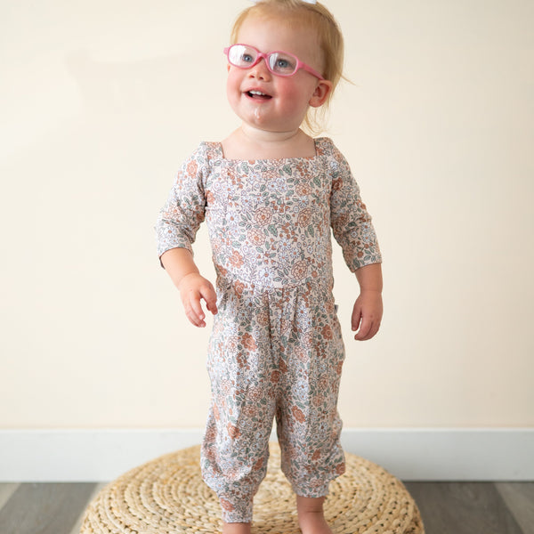 A toddler girl wears a floral print romper and pink glasses.