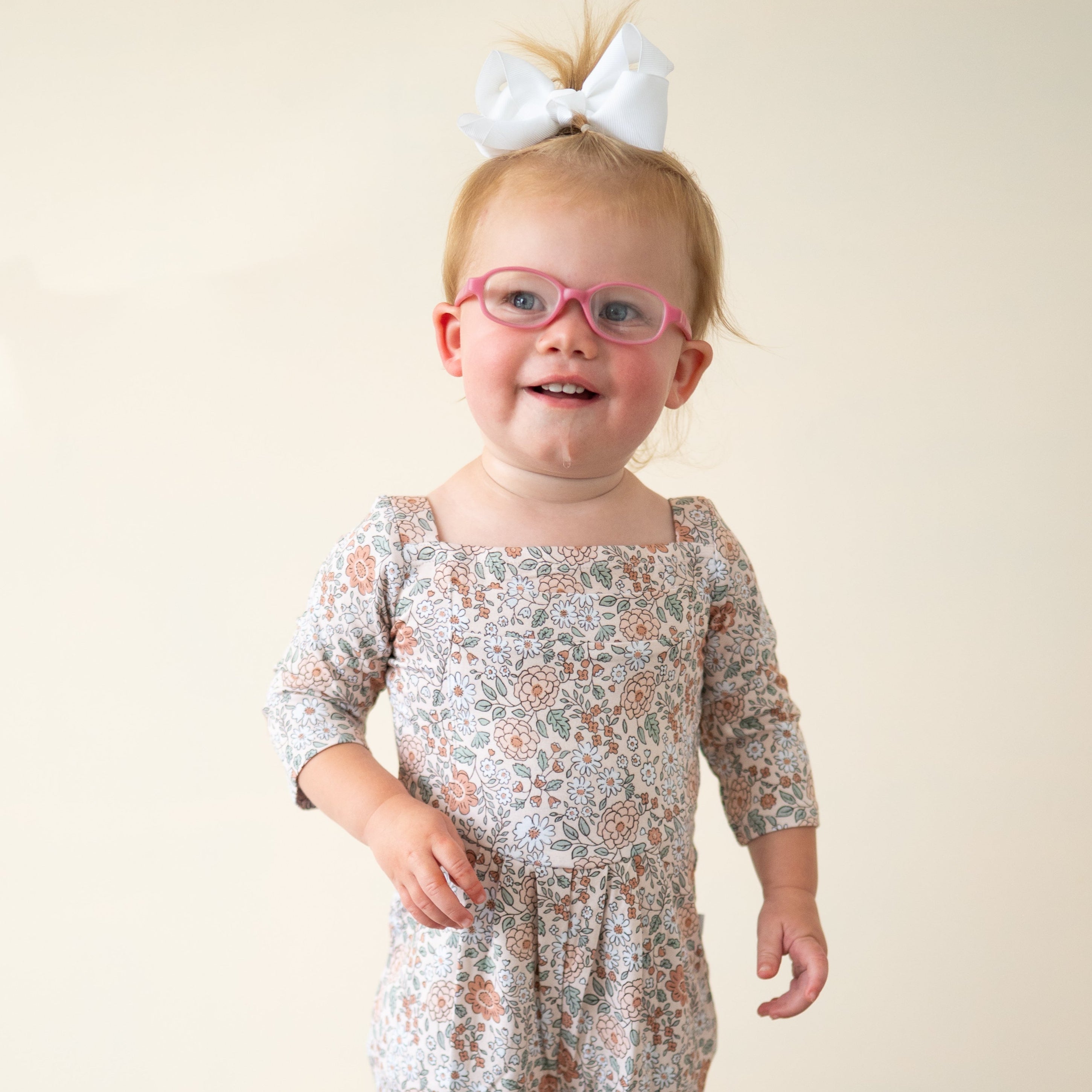 A toddler girl wearing a floral romper and pink glasses smiles.